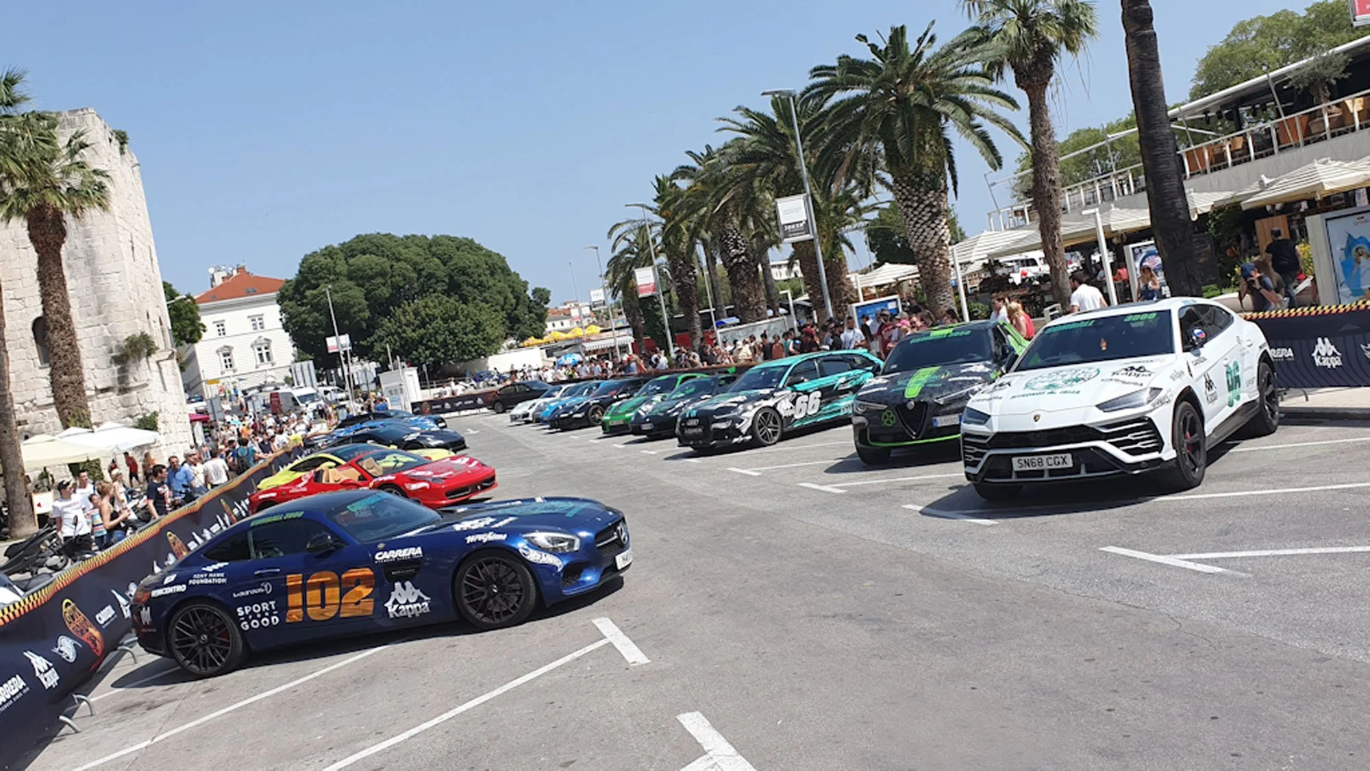 A lineup of racing cars parked along a street with an audience and palm trees in the background