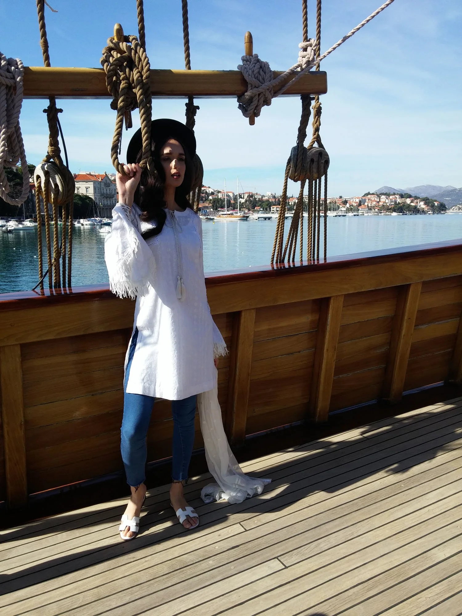 A woman standing on a wooden deck near a ship with ropes and wooden railings, with a harbor and buildings in the background under a clear sky.