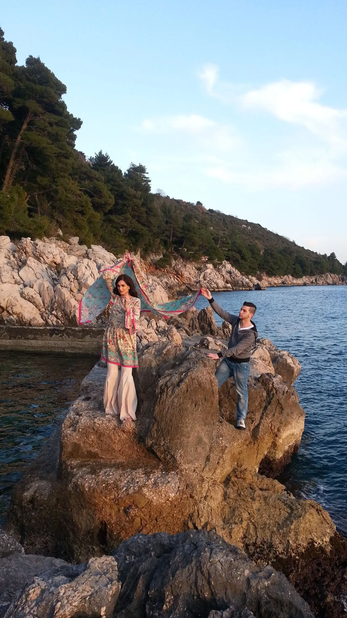 A man and a woman standing on large rocks by the water, holding a colorful scarf between them with a forested hillside in the background during sunset.