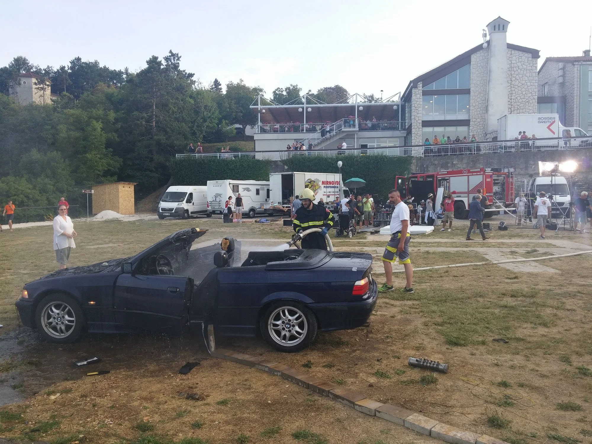 Scene of an outdoor event with a black convertible car in the foreground, a group of people, some in uniforms, and emergency vehicles, with a building and trees in the background.