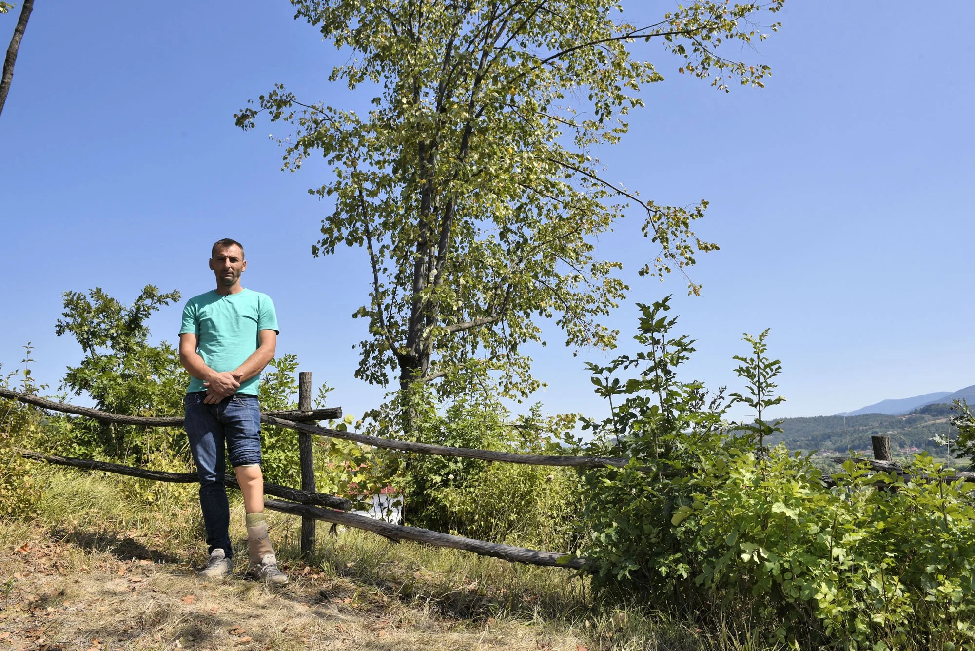 A man standing outdoors near a wooden fence with green trees and hills in the background, under a clear blue sky.