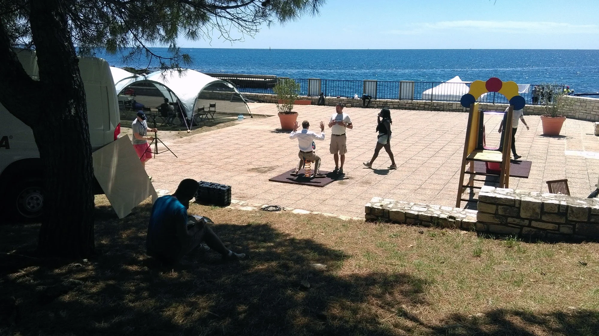 People working on a photoshoot near a waterfront, with some individuals posing, a photographer, and vintage equipment, under trees with the ocean in the background.