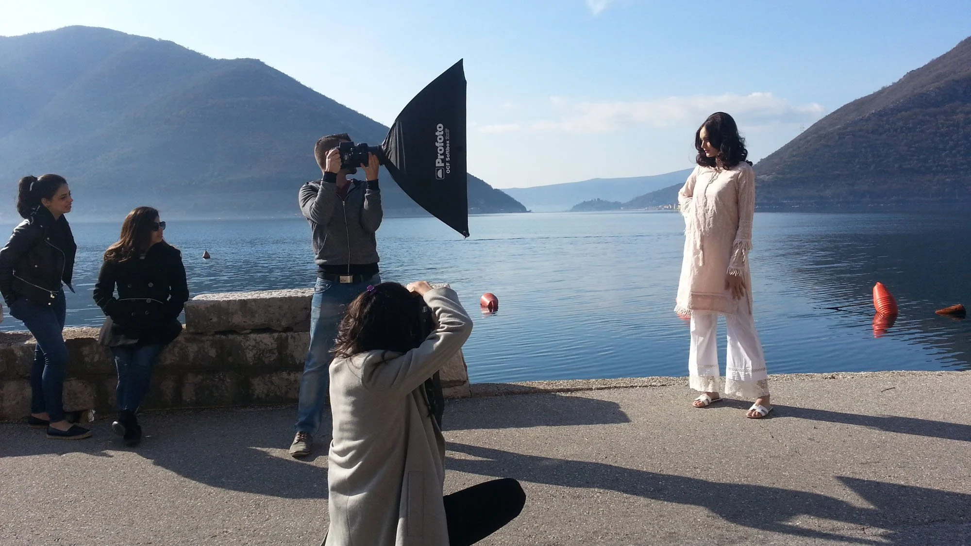 A woman in a cream-colored dress standing by a lake with mountains in the background, while a photographer and two women nearby are taking pictures of her on a sunny day.