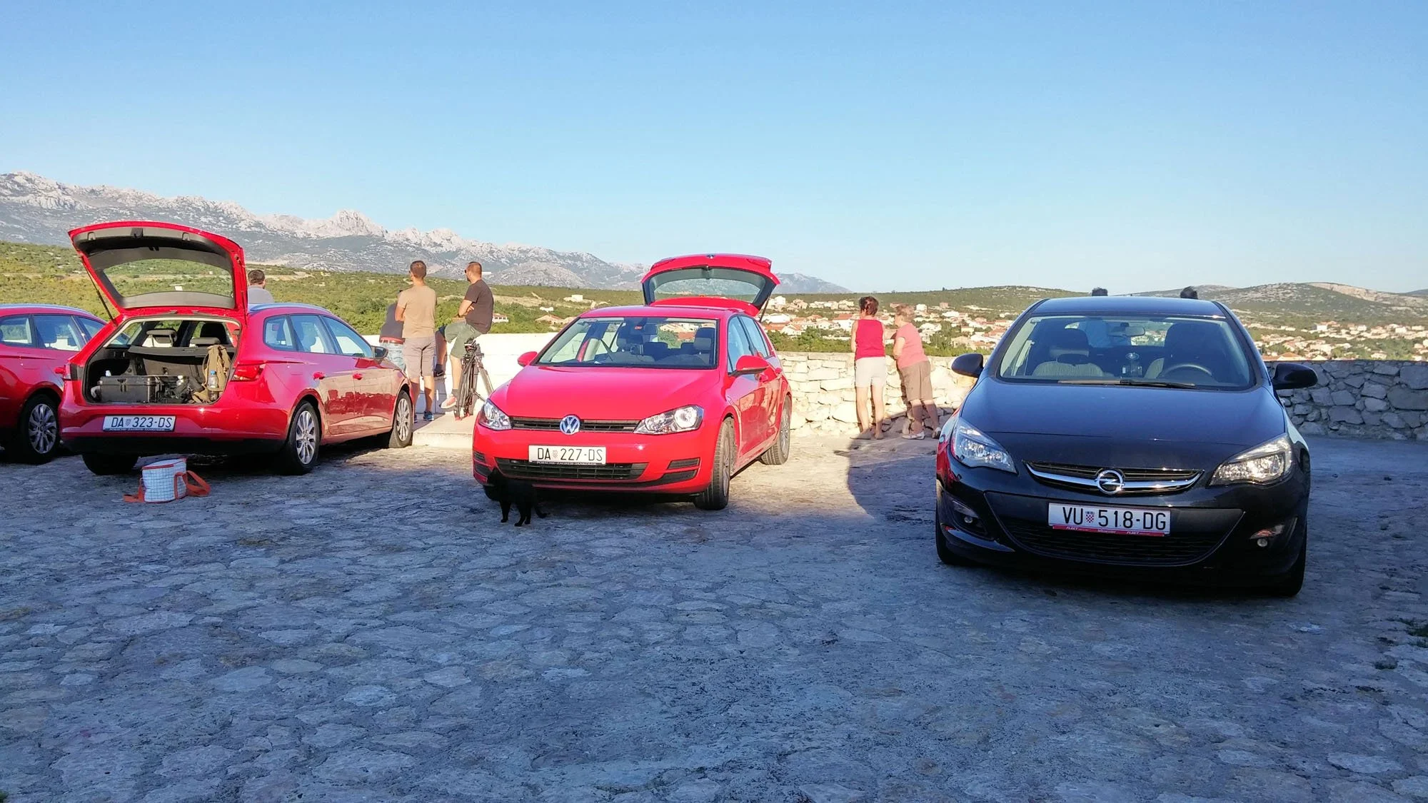Several cars parked outdoors with their trunks open and a group of people standing around, in a scenic mountain area under a clear blue sky.