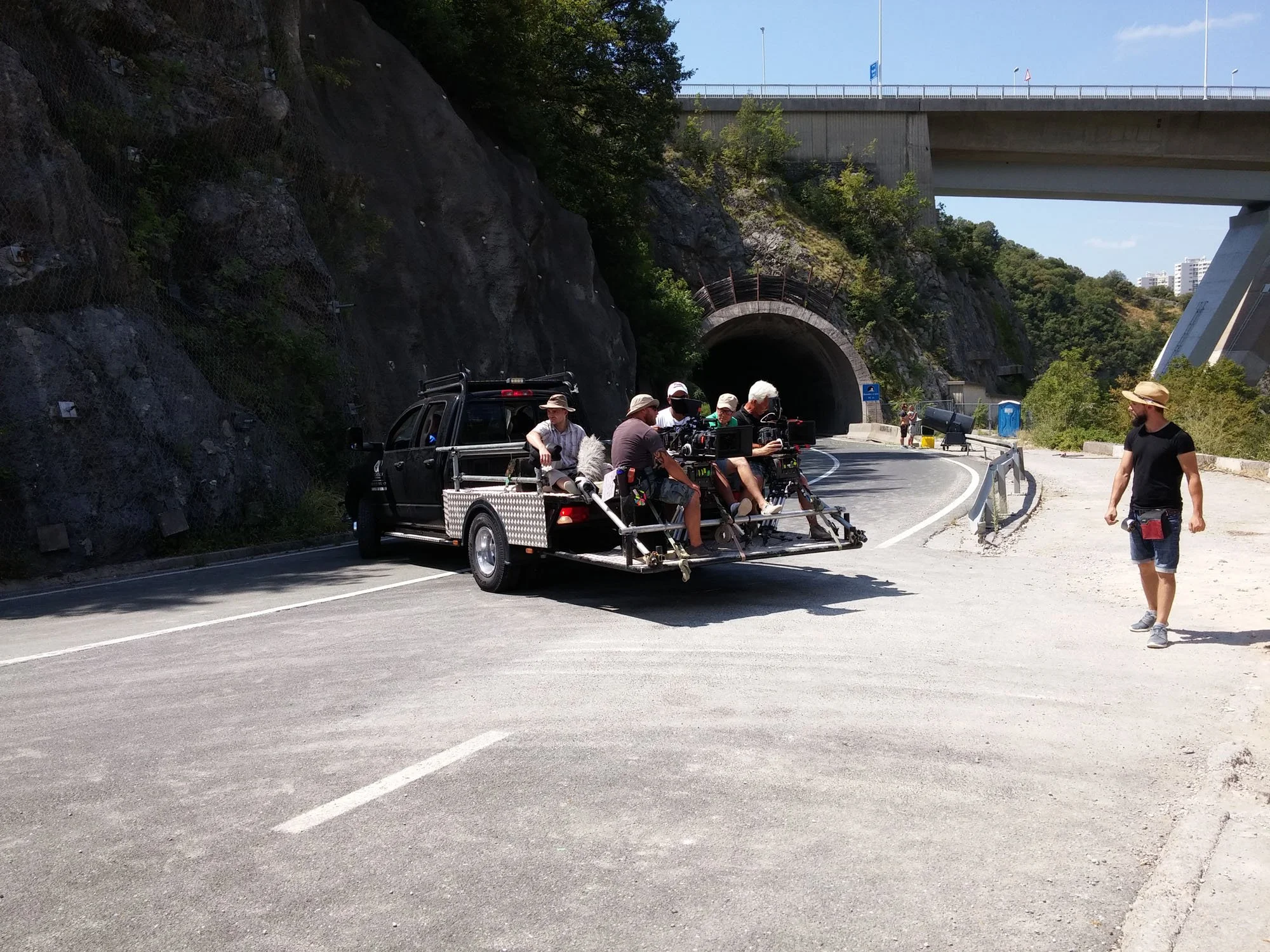A film crew is filming on a mountain road, with equipment and crew members sitting on a truck trailer, under a bridge with a tunnel in the background.