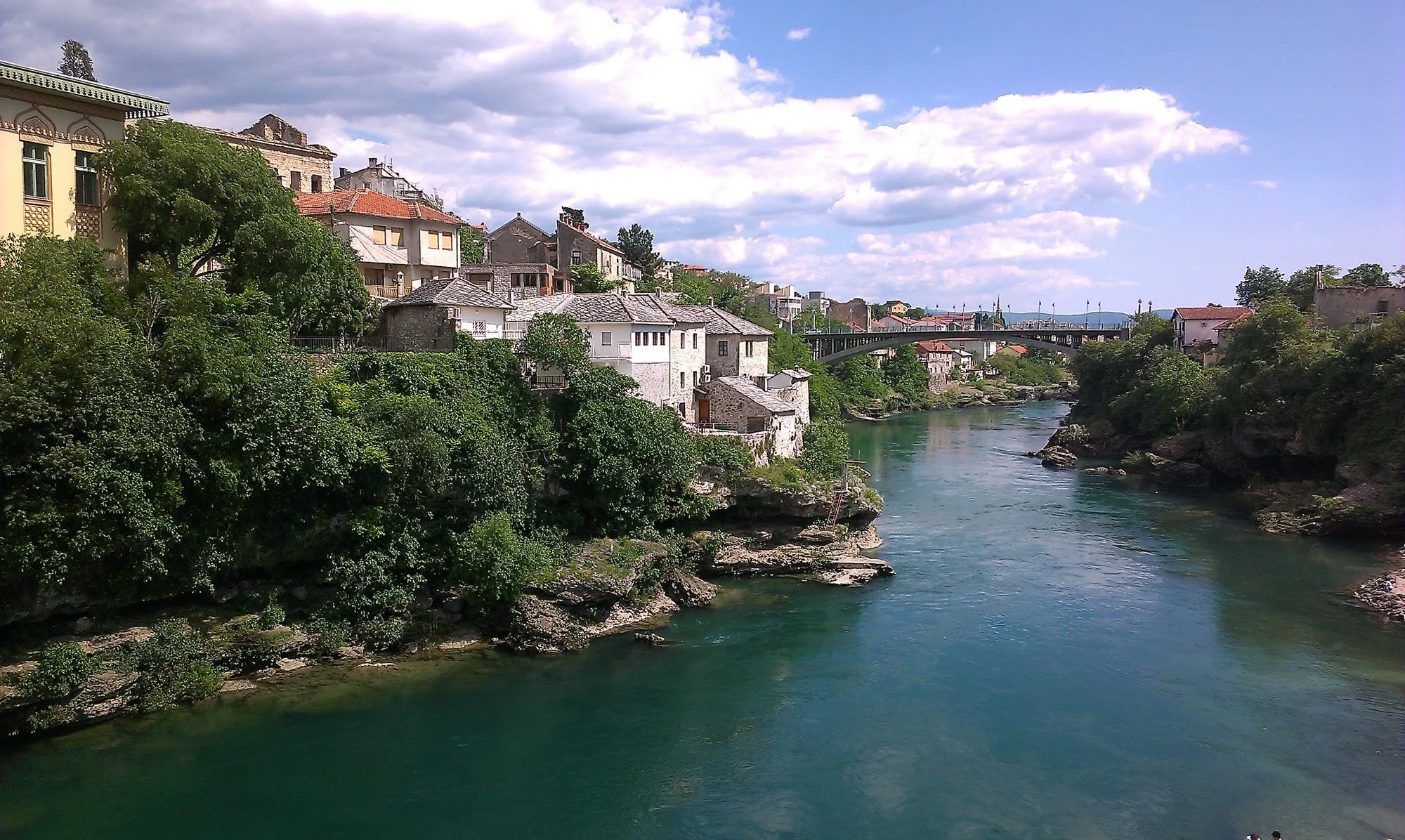 A river flowing through a town with houses on a hillside, a bridge crossing the river, under a partly cloudy sky.