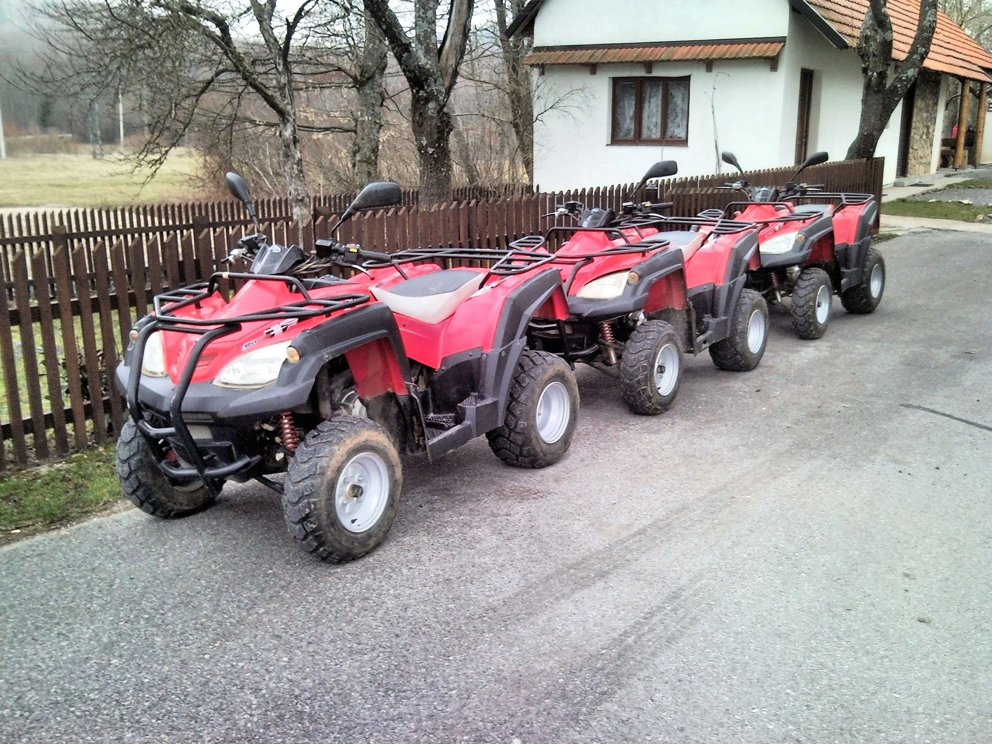 A row of four red all-terrain vehicles (ATVs) parked on a gravel driveway, lined up facing forward, with a fenced yard, trees, and a white house with a small porch in the background.