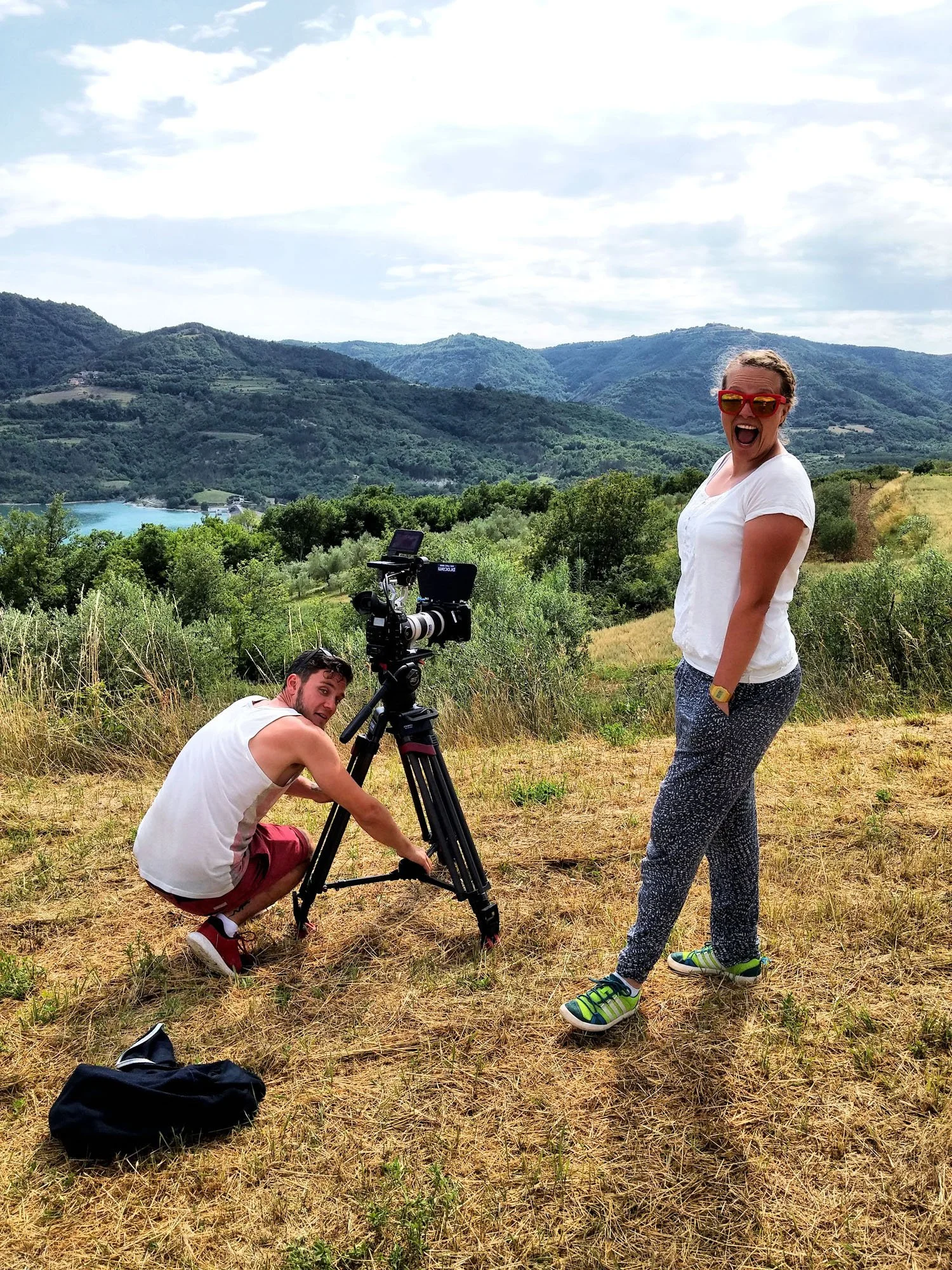 A woman with red sunglasses, white shirt, and patterned pants standing outdoors on a grassy hill, smiling with her mouth open. A man with a white sleeveless shirt and red shorts crouching down adjusting camera equipment near her. The background featu