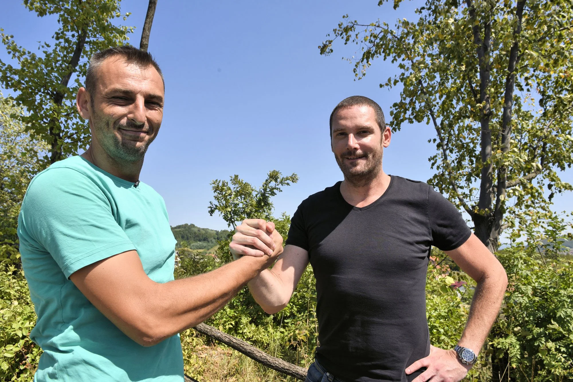 Two men are shaking hands outdoors under a clear blue sky, surrounded by trees and greenery.