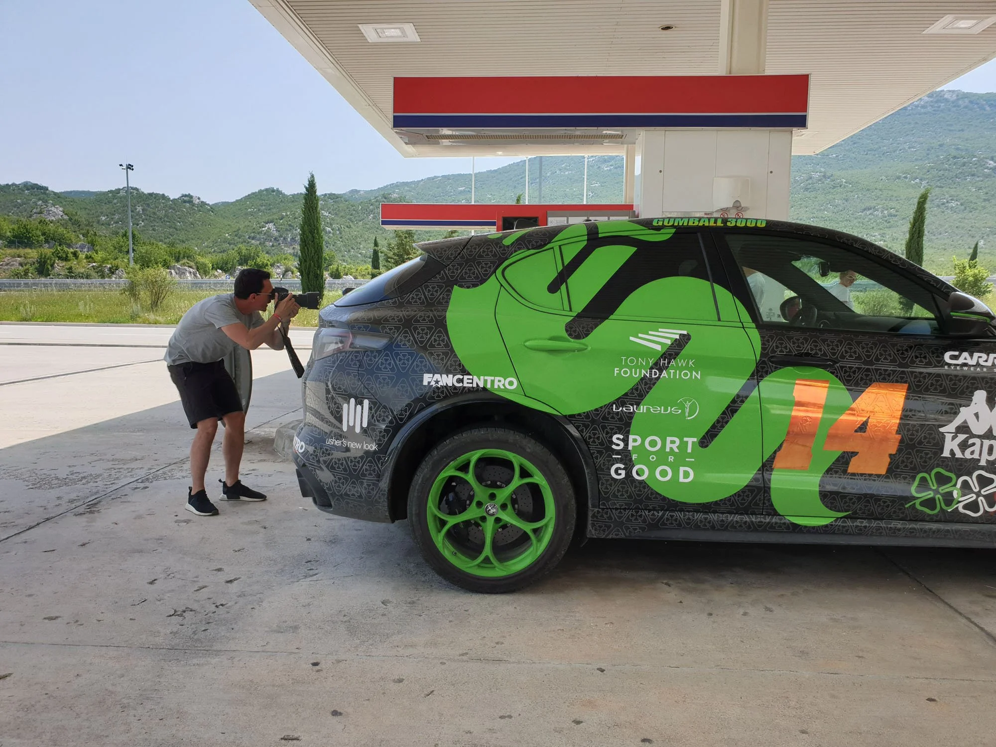 A man takes a photograph of a race car at a gas station under a canopy, with green hills in the background. The race car is green and black with sponsors and a large number 44 on the side.