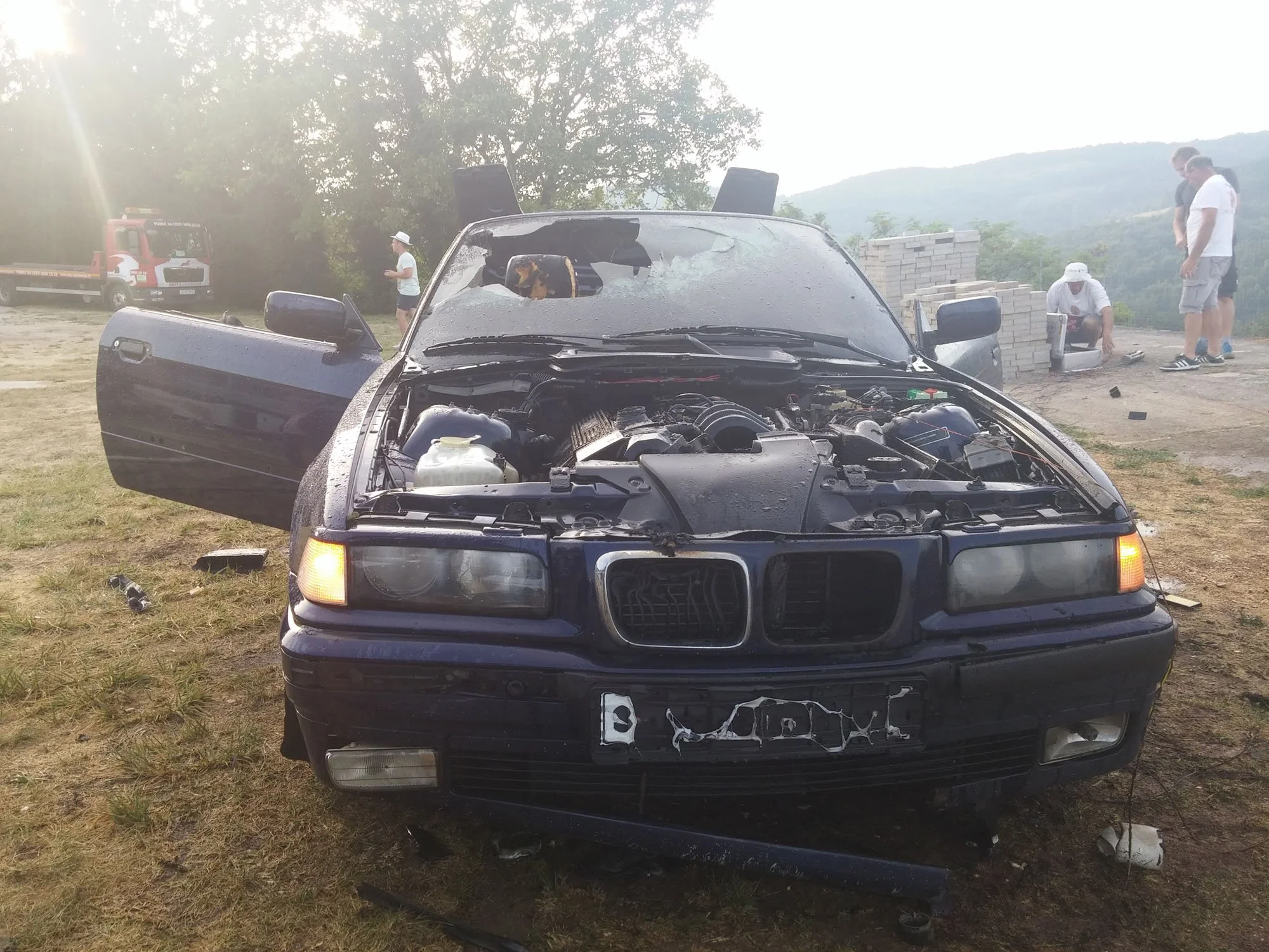A black car with a severely damaged front hood and broken windshield, parked outdoors on grass, with debris and broken parts around it. In the background, several people are near a brick wall, with some inspecting or working on repairs, and a fire tr