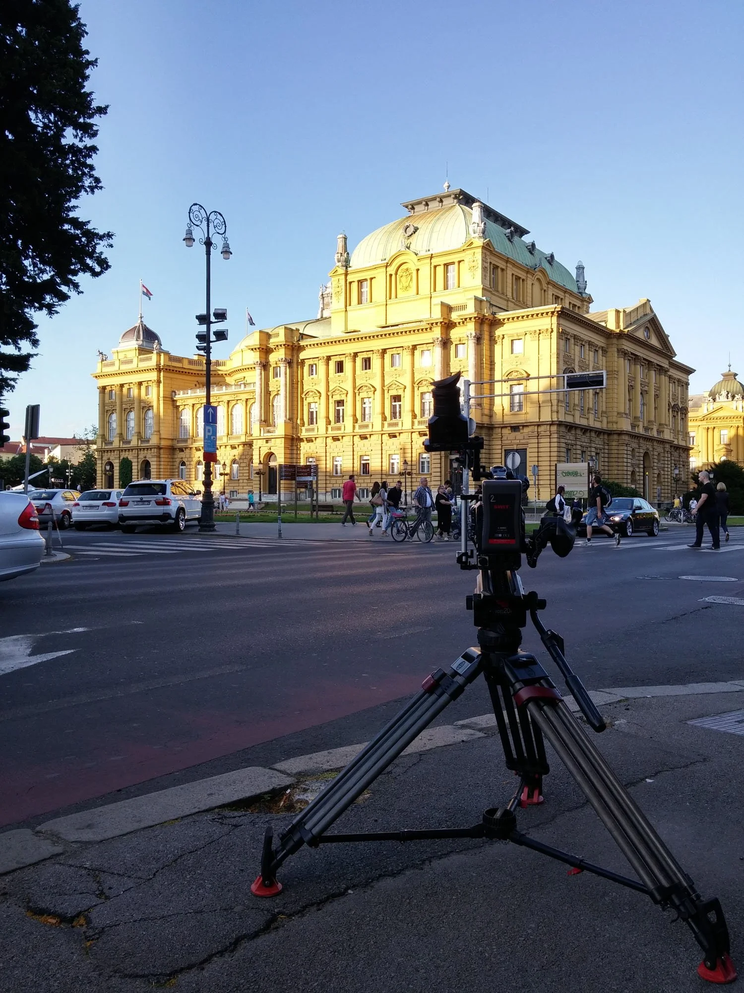A historic yellow building with ornate architecture under a clear blue sky, with a street scene including parked cars, pedestrians, bicycles, street lamps, and a tripod camera in the foreground.