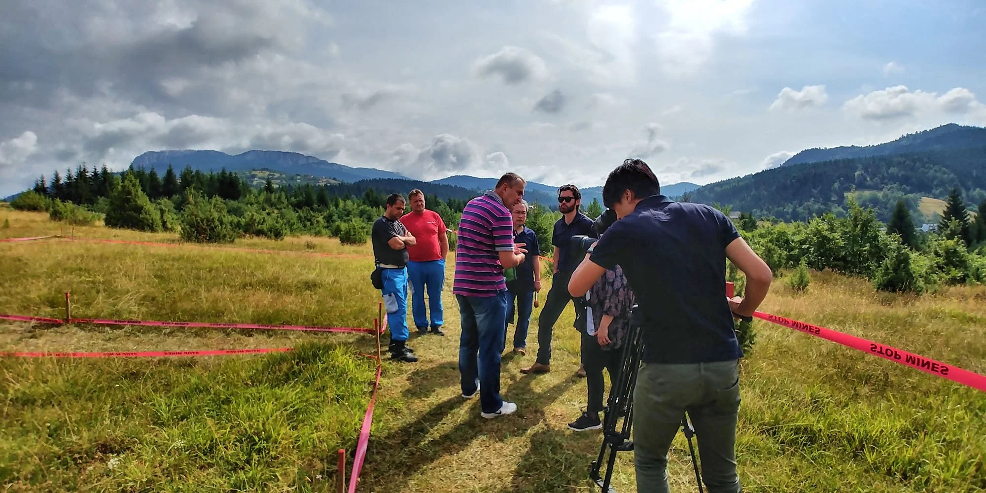 Group of people standing on a field with mountains and trees in the background, some talking and others observing, with red caution tape on the ground.