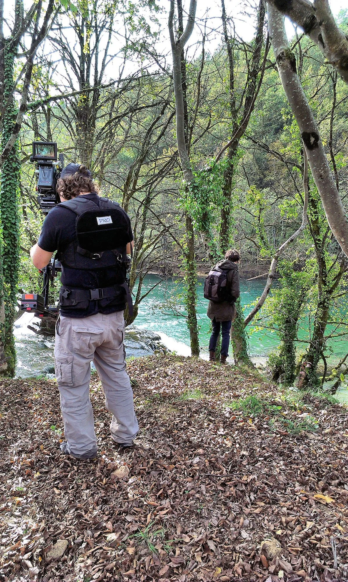 A person with curly hair wearing a black shirt, beige pants, and a camera backpack takes photos of another person standing near a river in a wooded area, surrounded by trees and fallen leaves.