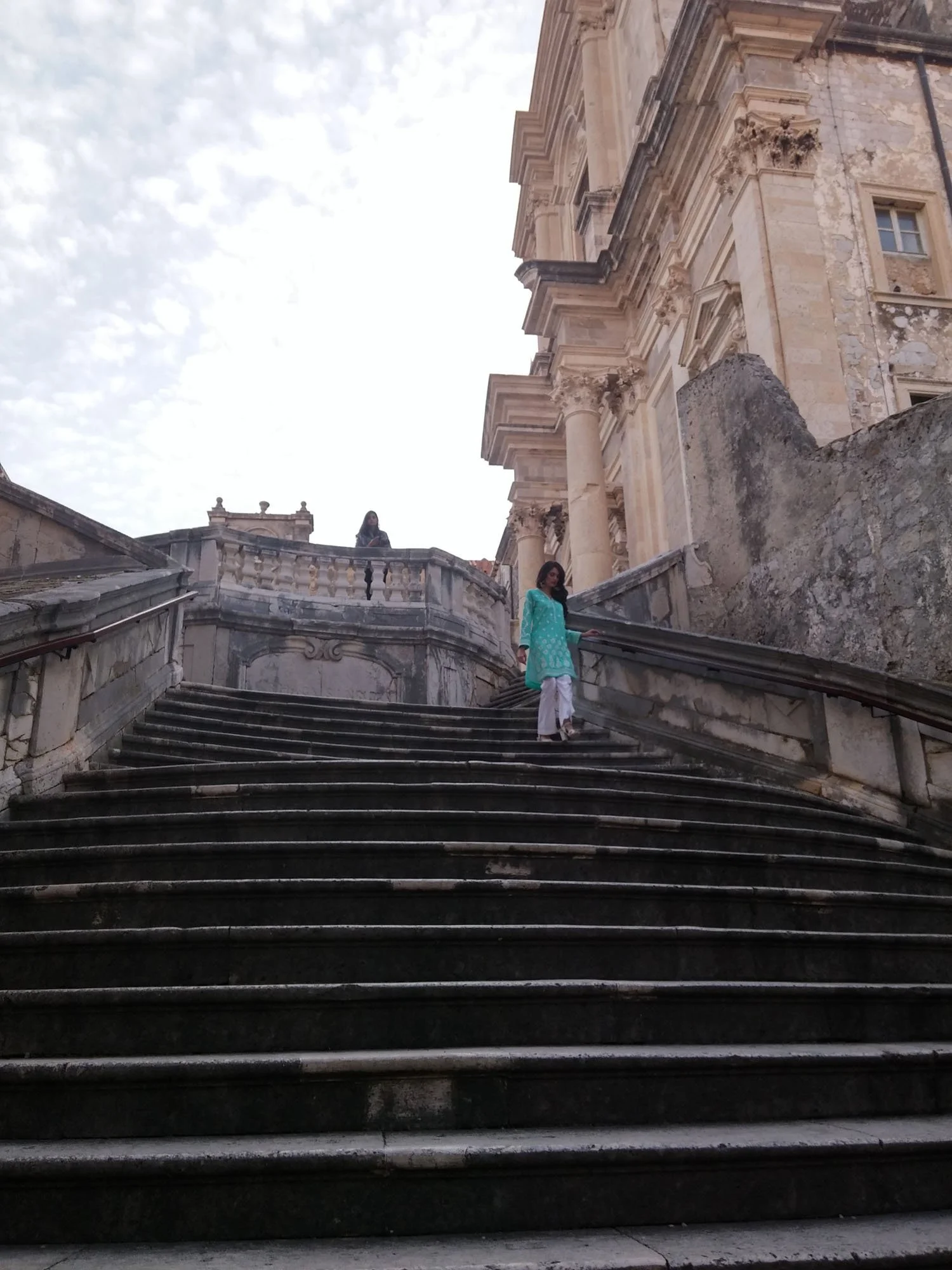 The image shows a set of stone stairs leading up to an ornate, historical building made of stone with columns and detailed architectural features. Two women, one in a traditional turquoise outfit and the other in darker clothing, are descending the s