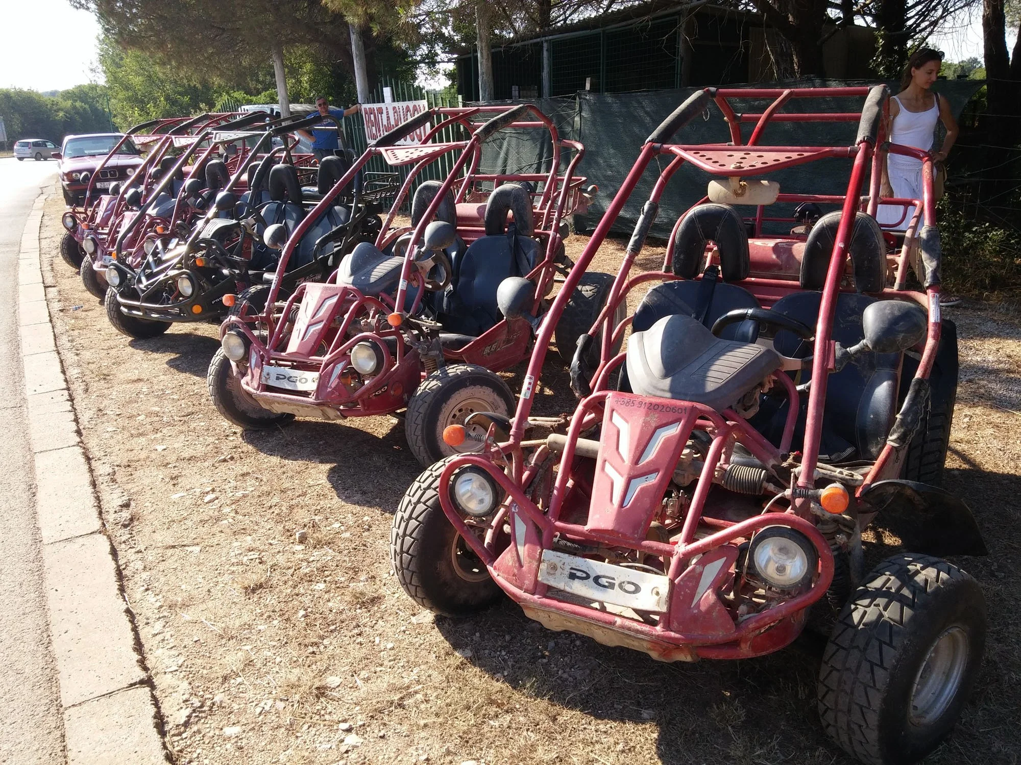 Row of pink and black pedal go-karts parked on dirt ground along a sidewalk, with two women nearby, one standing and one sitting, and trees and cars in the background.