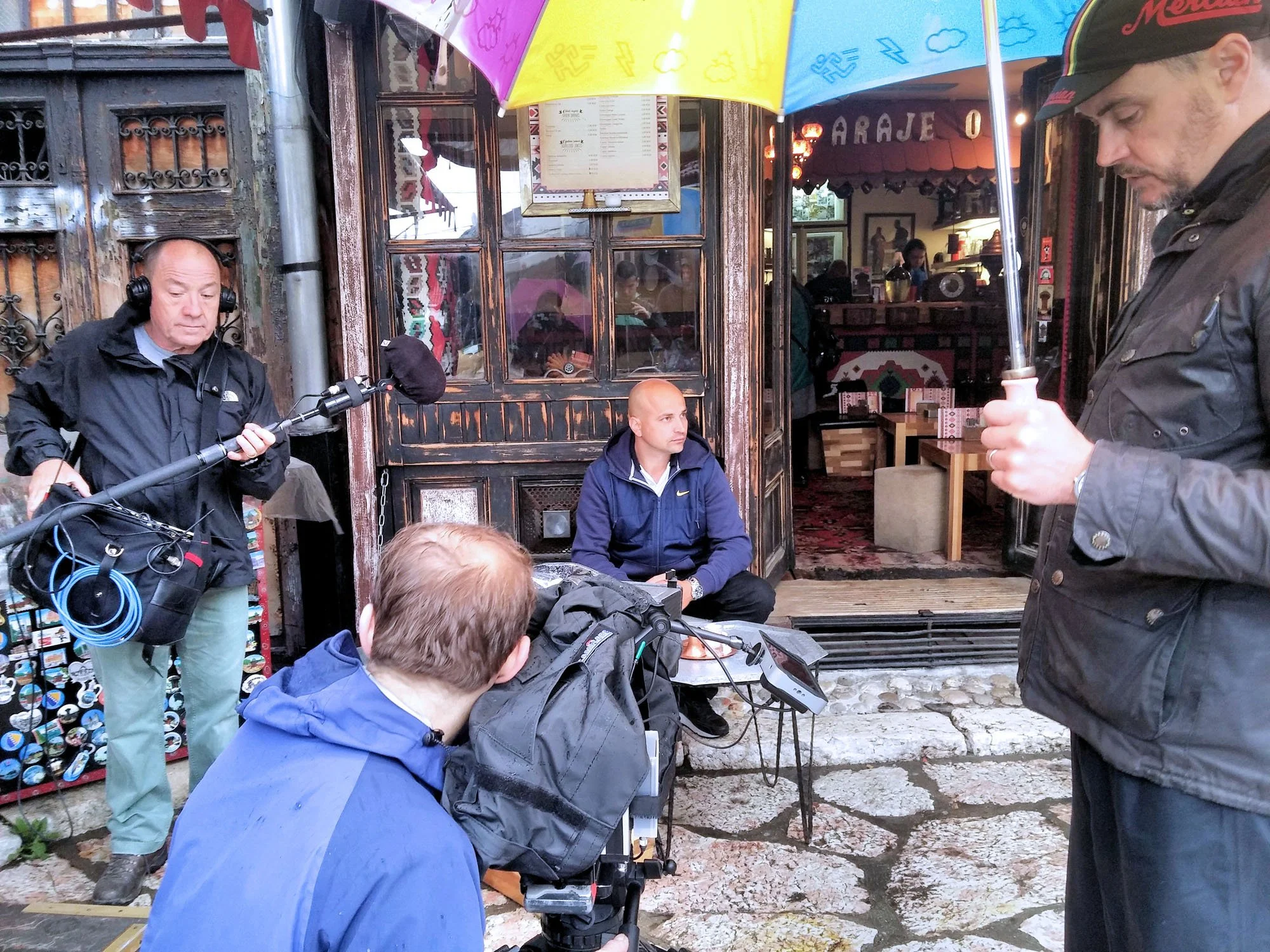 A behind-the-scenes shot of a film or video production outside a restaurant or cafe. Several crew members are operating equipment, with one person holding a boom microphone and another operating a camera. A man with a jacket and cap is in the foregro