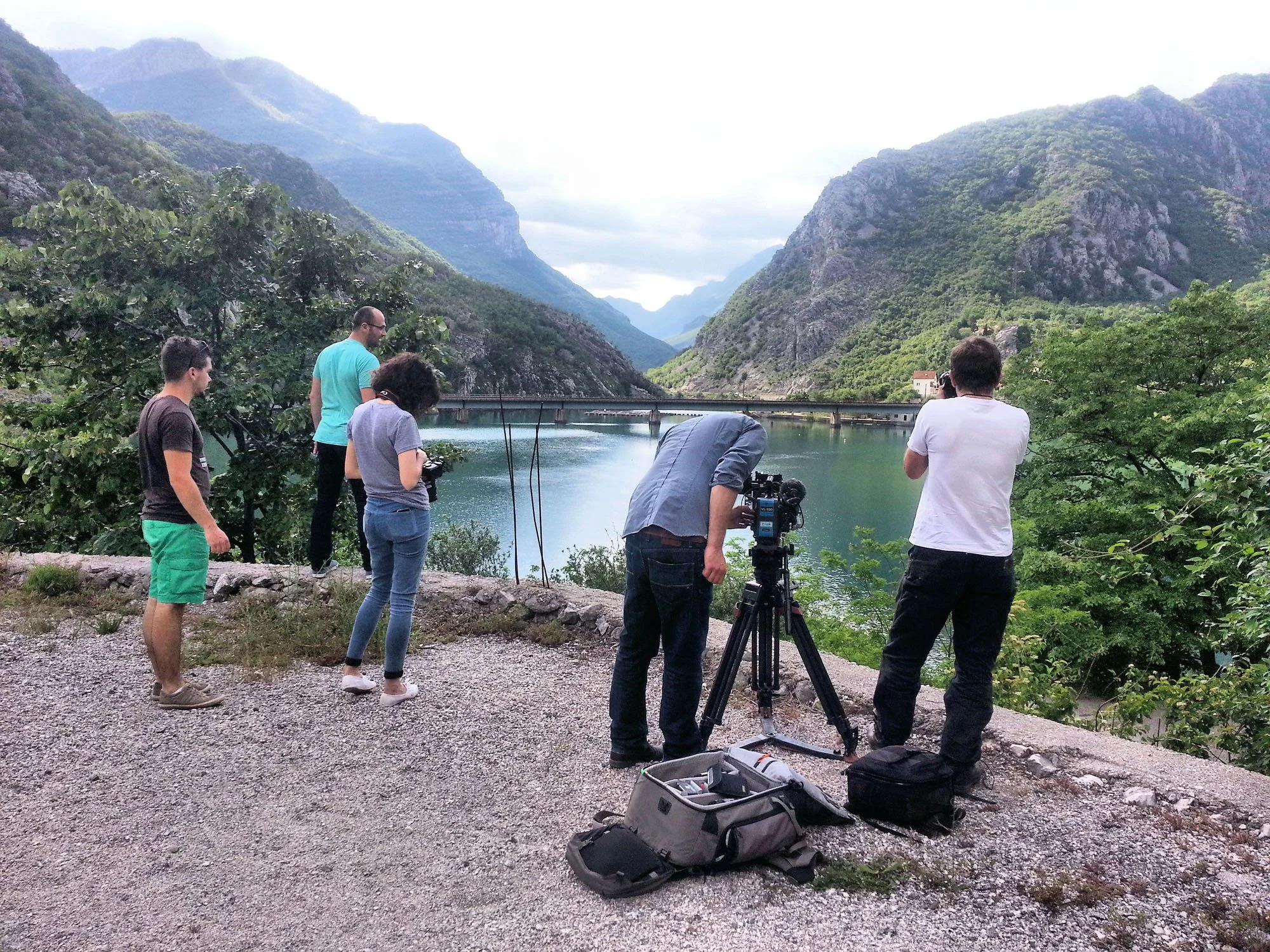 Group of people setting up camera equipment on a gravel path by a serene lake surrounded by green mountains under a cloudy sky.
