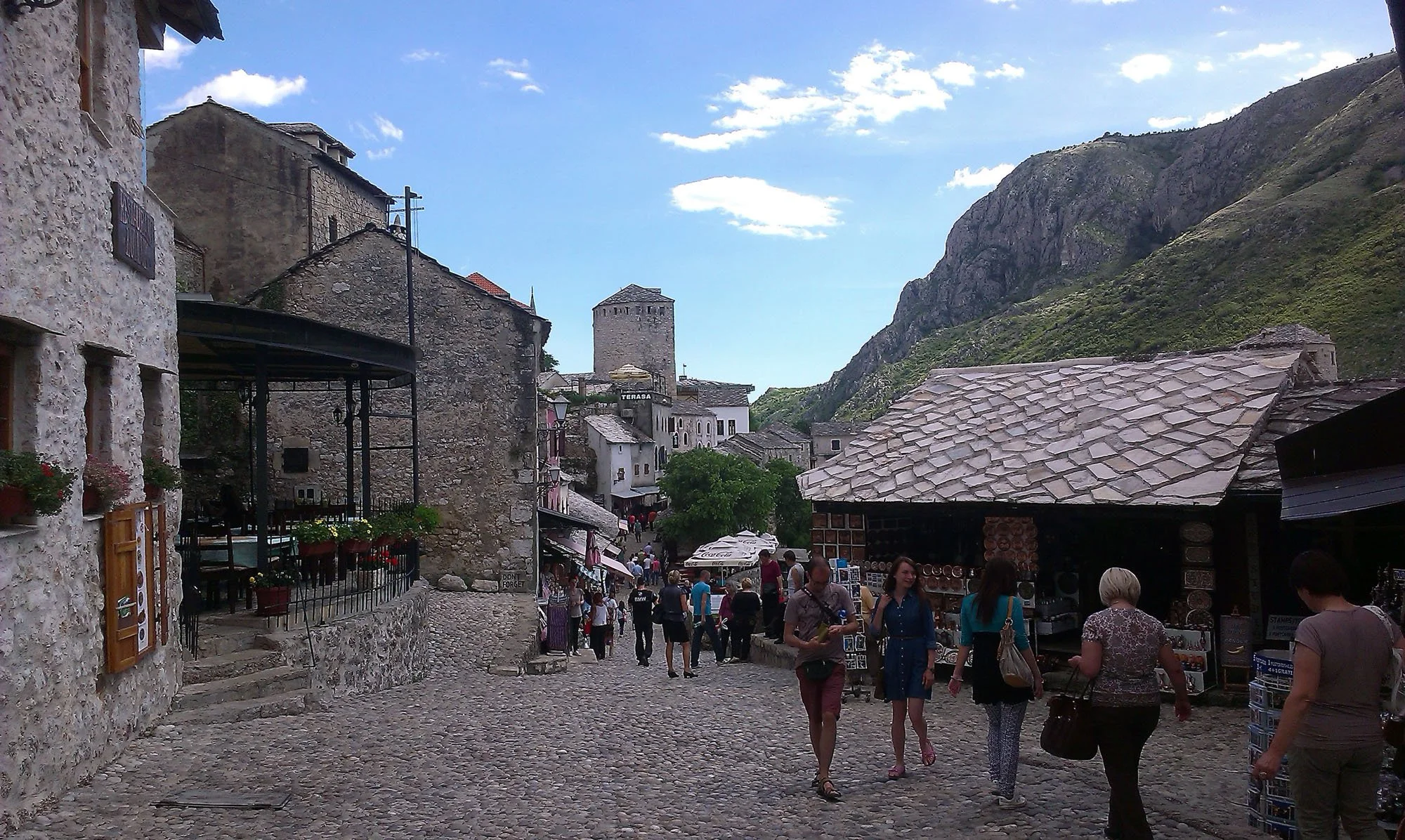A busy street in a mountain village with stone buildings, a cobblestone path, and people walking around, with mountains and blue sky in the background.