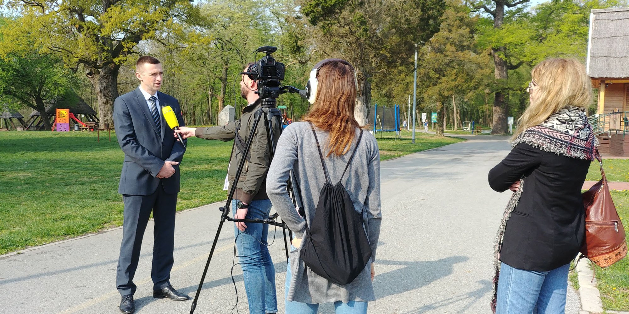 A man in a suit giving an interview to a woman with a microphone at an outdoor park with trees, a path, and a wooden building in the background.