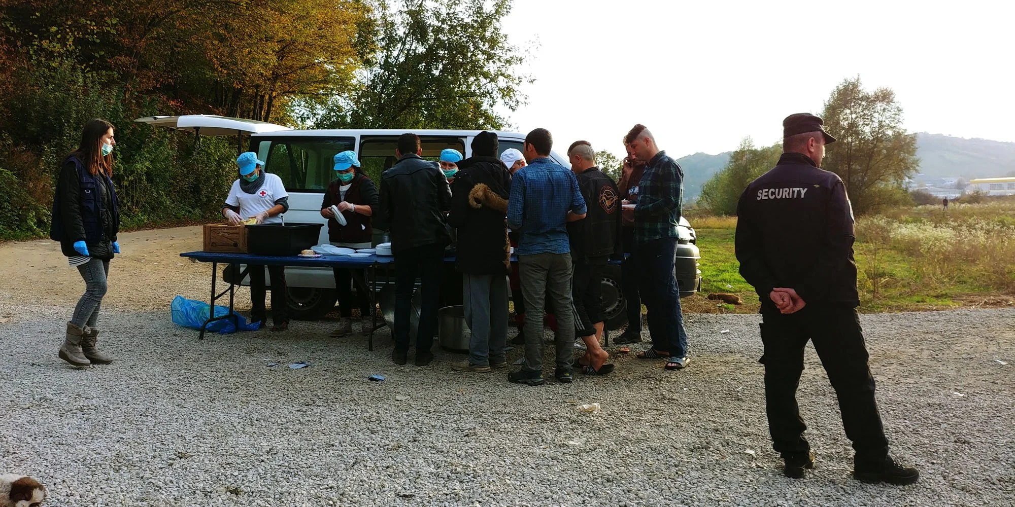 A group of people, including medical personnel and security personnel, gathered around a food or supply table near an open vehicle on a gravel path in an outdoor setting with trees and hills in the background.