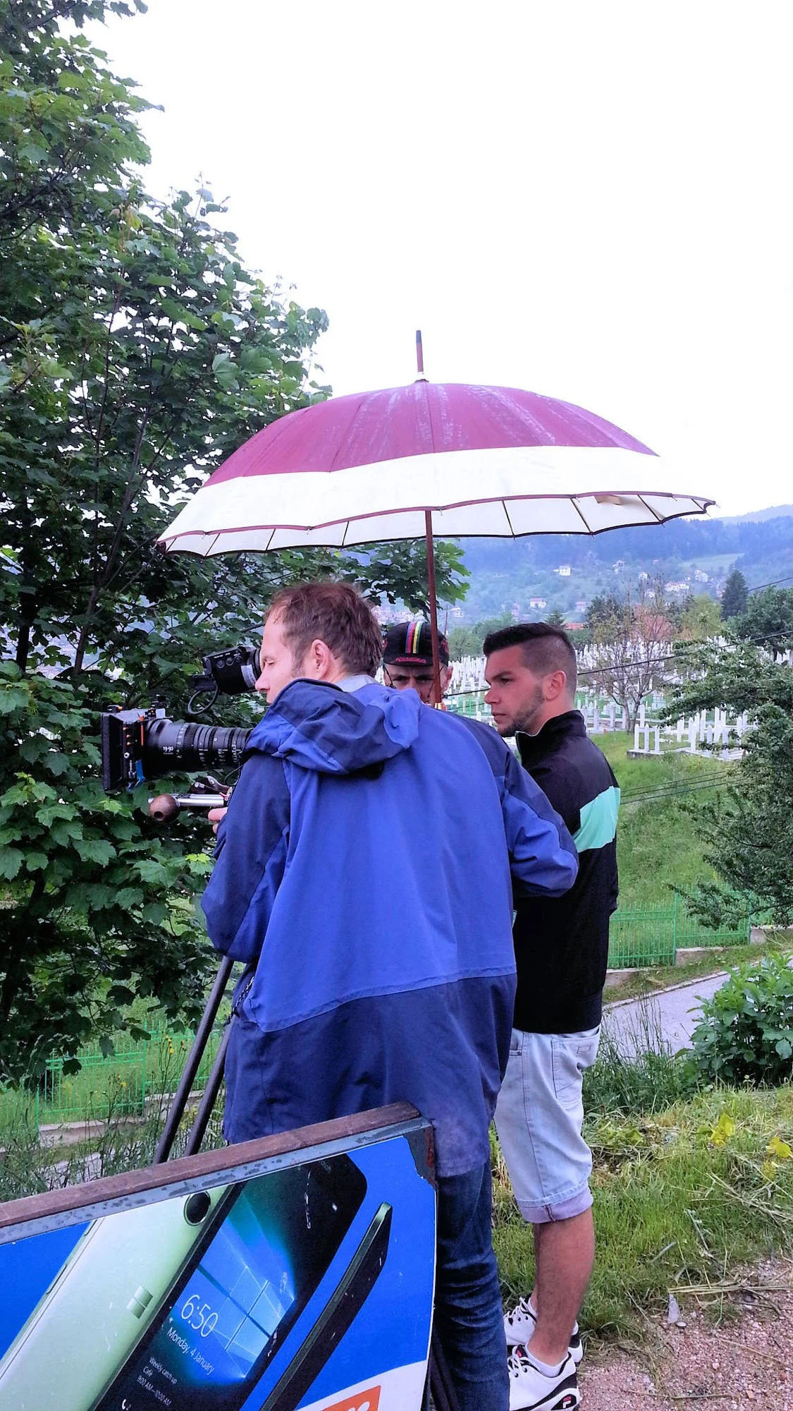 Three men outdoors with a pink and white umbrella, one filming with a camera, another person is behind him, and a third man standing in front of the camera with arms crossed. There are trees and a hilly landscape in the background.