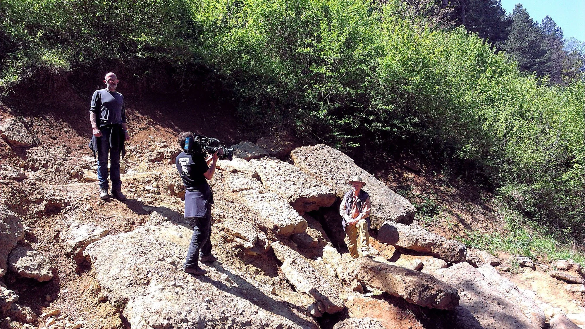 A group of four people, including a man sitting on rocks and a woman filming with a video camera, are exploring a rocky outdoor area surrounded by green trees under a clear blue sky.