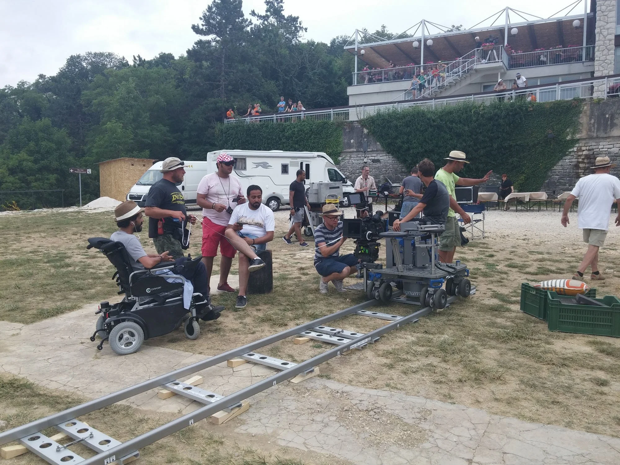 Filmmaking crew on a set with a camera crane, people, and equipment outdoors near a building with stairs and balcony.