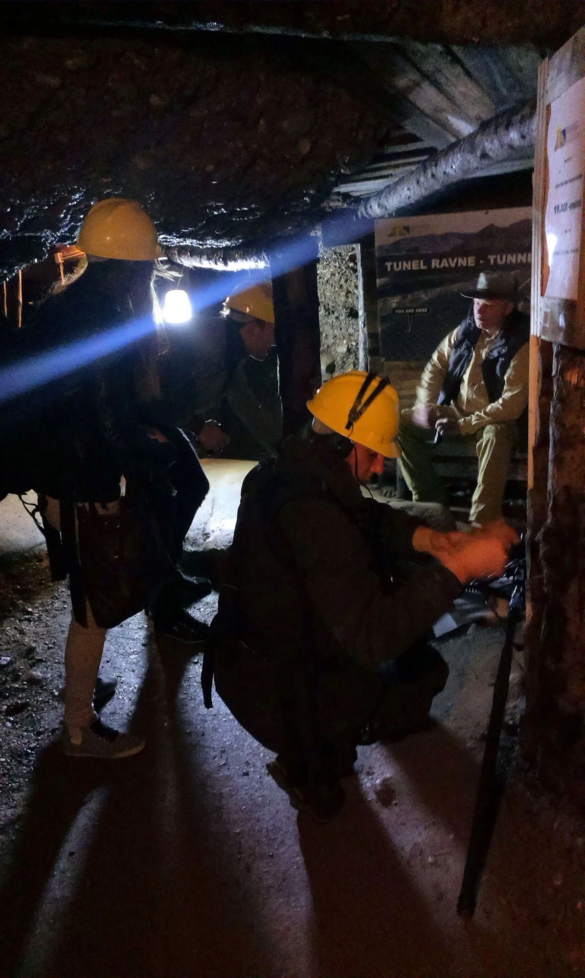 A group of people wearing yellow mining helmets and outdoor clothing inside a dark tunnel, with one person sitting and another standing, possibly inspecting or working on equipment.