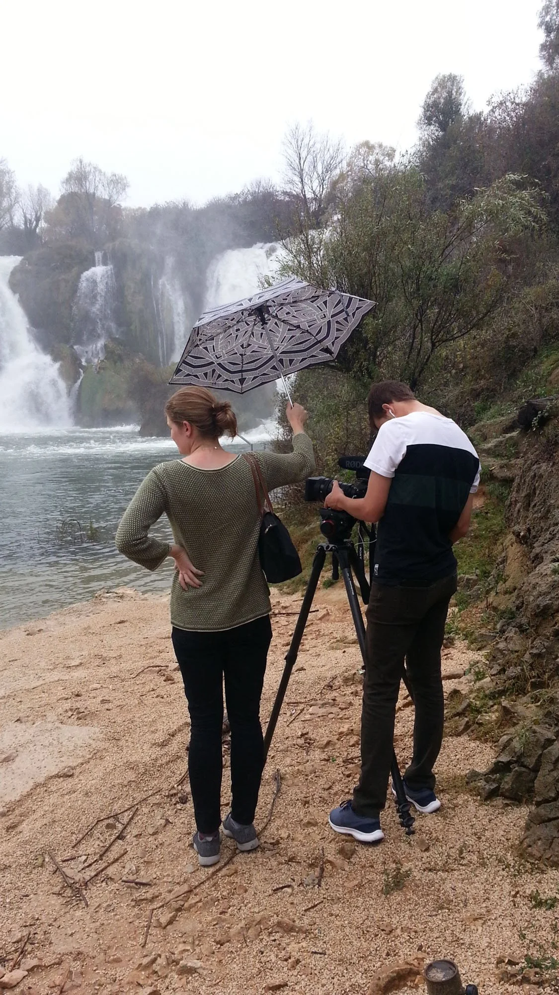 Two people stand on a rocky riverbank with a waterfall in the background. One person holds a patterned umbrella, and the other is looking at a camera mounted on a tripod.