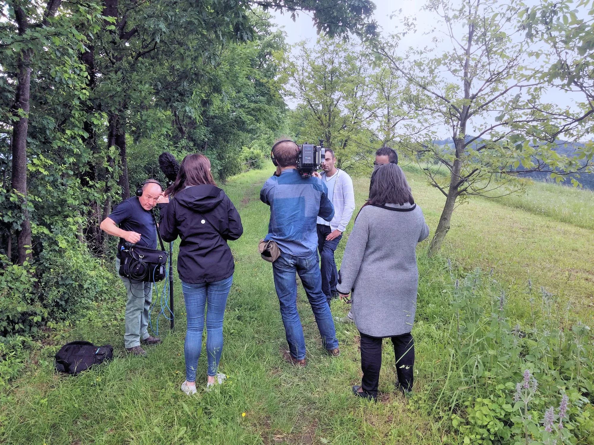 A group of people, including a man with headphones and a camera, are outdoors on a grassy path surrounded by trees. They appear to be filming or conducting an interview.
