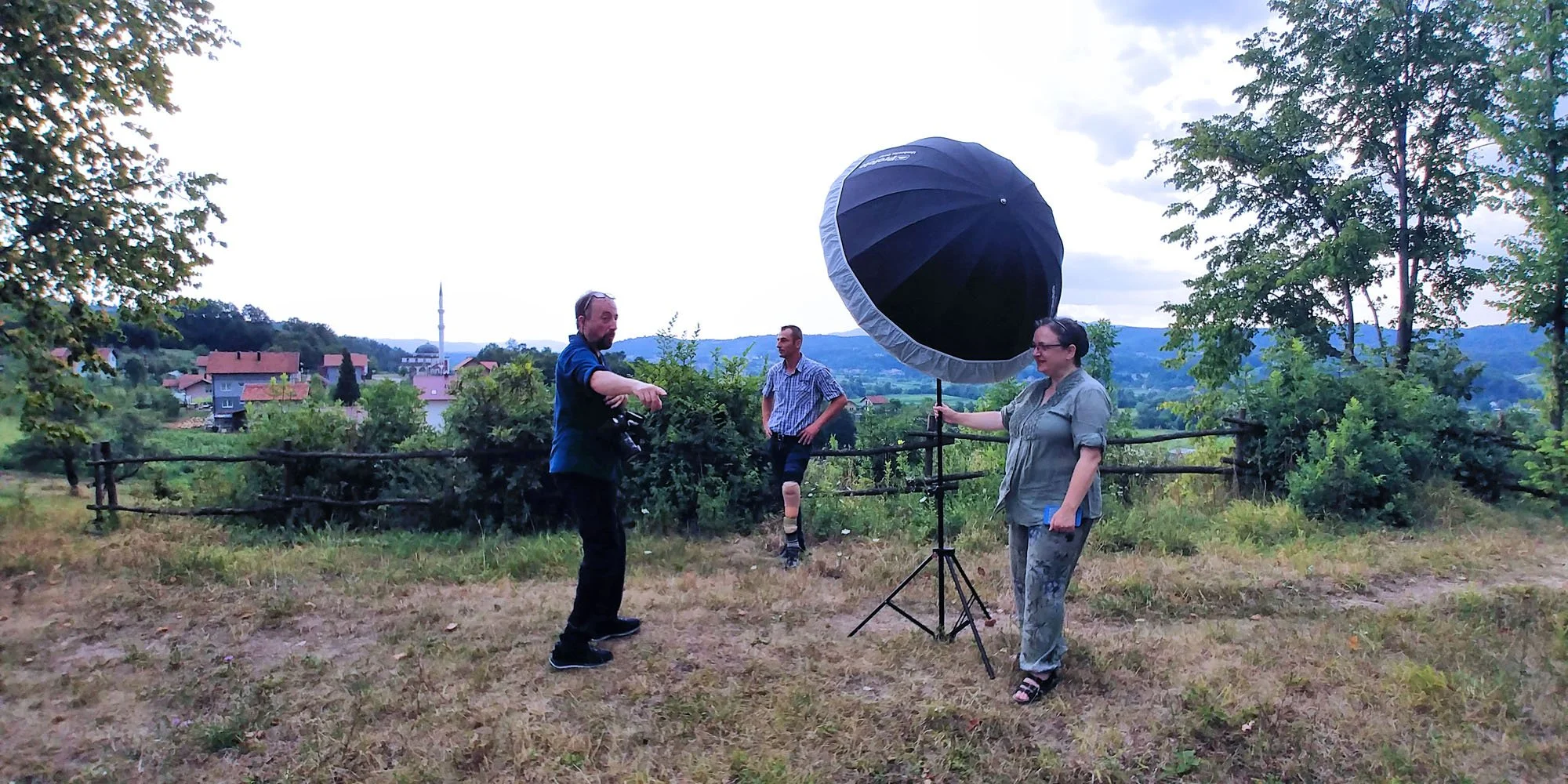 Three people setting up photography equipment outdoors in a rural area, with a large umbrella reflector and a camera on a tripod, and a scenic view of houses, trees, and distant hills in the background.