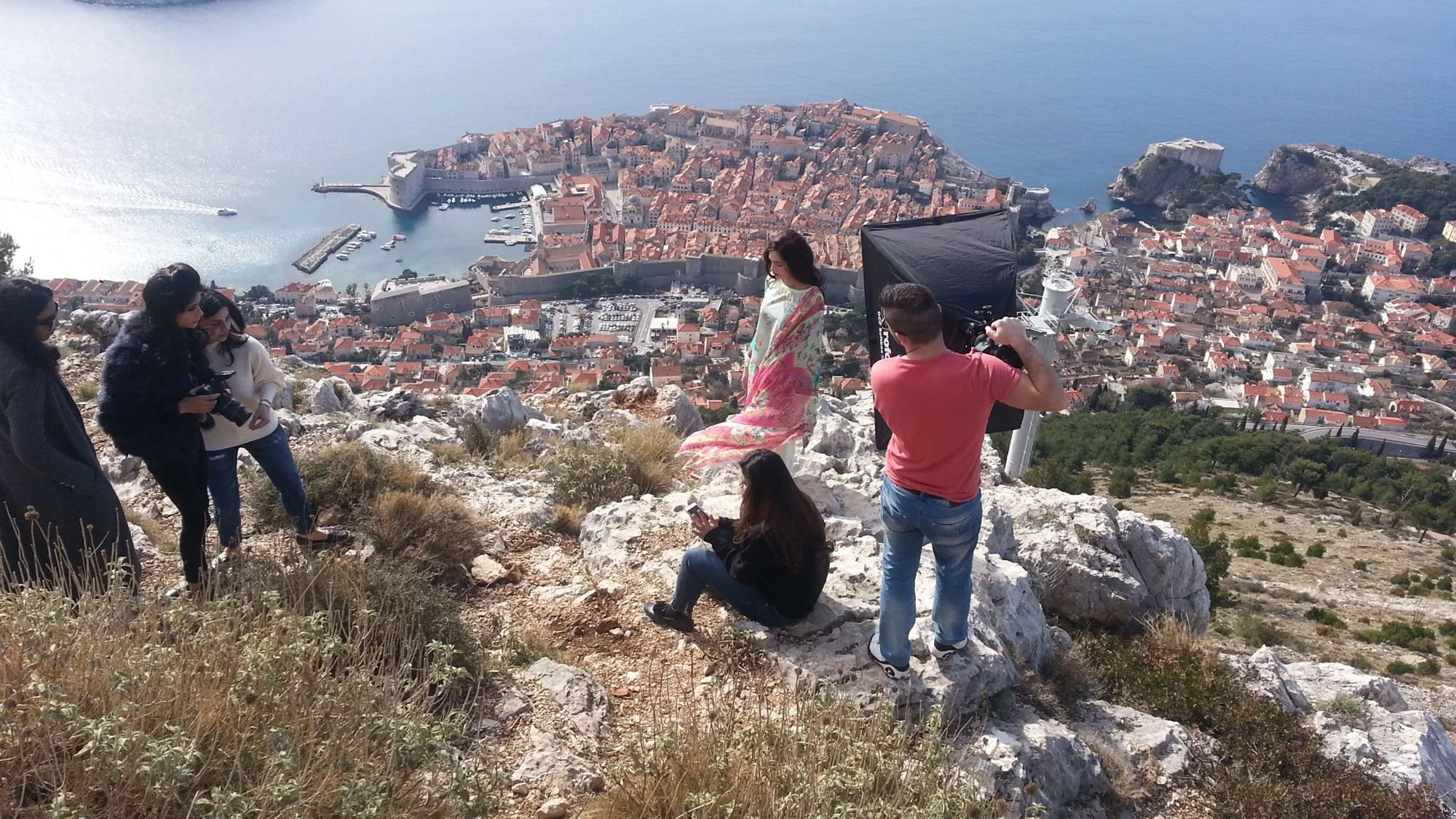A photoshoot scene on a rocky hillside overlooking a historic coastal city with red-roofed buildings and a harbor, featuring a woman in a floral dress being photographed by a man with lighting equipment, and several people nearby, some using smartpho