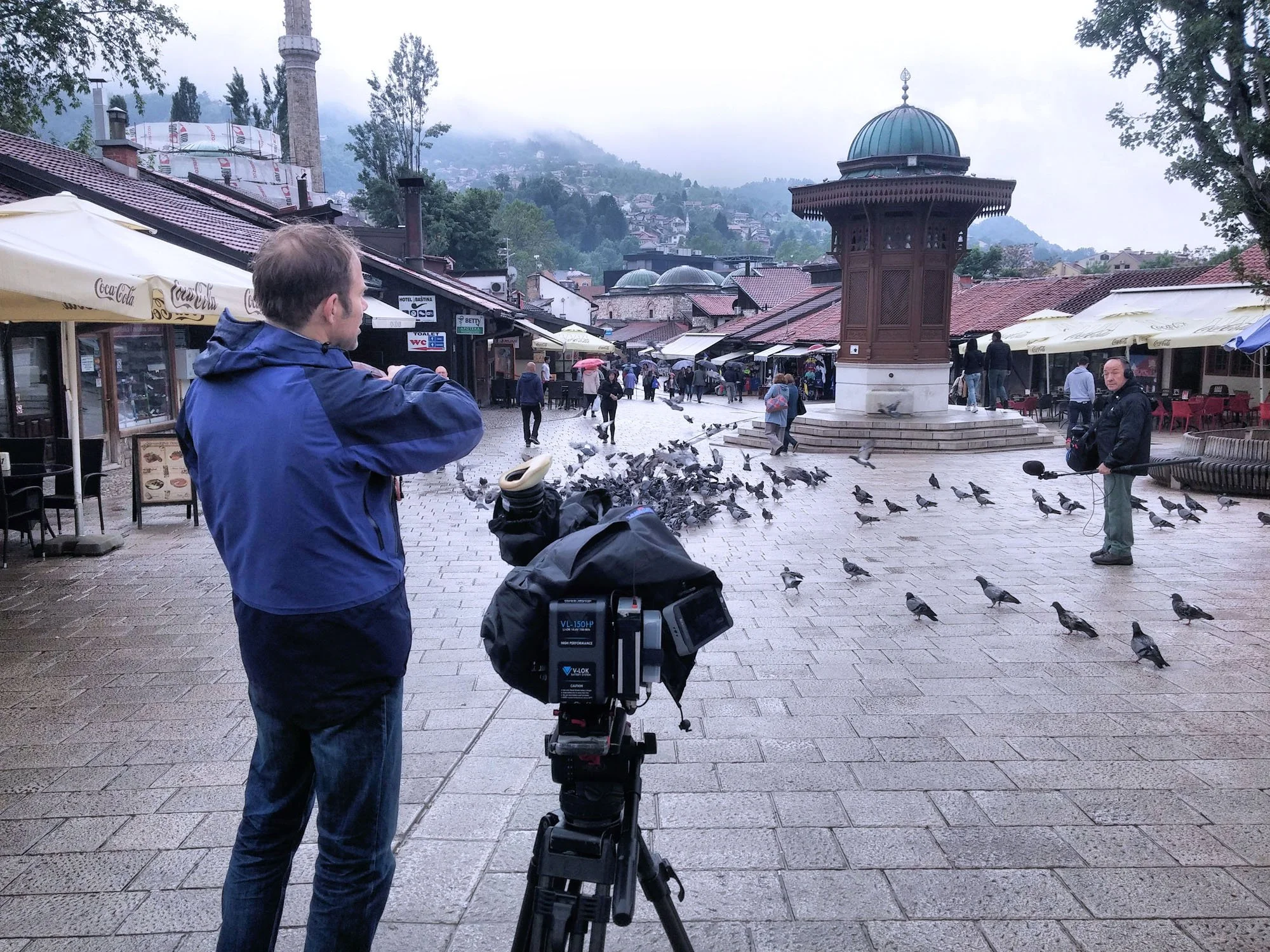 A man standing next to a camera on a tripod captures a scene in a crowded outdoor marketplace or town square, with pigeons on the ground and people walking. There are buildings with outdoor seating and a large wooden pavilion, with hills and mist in 