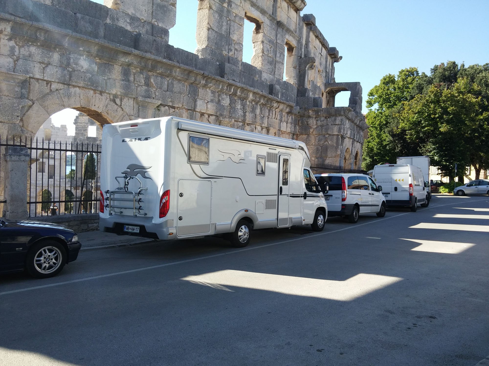 View of the ancient Roman amphitheater, the Colosseum, with part of its stone structure visible behind a row of parked cars and vans, and a green tree on the right, under a clear blue sky.