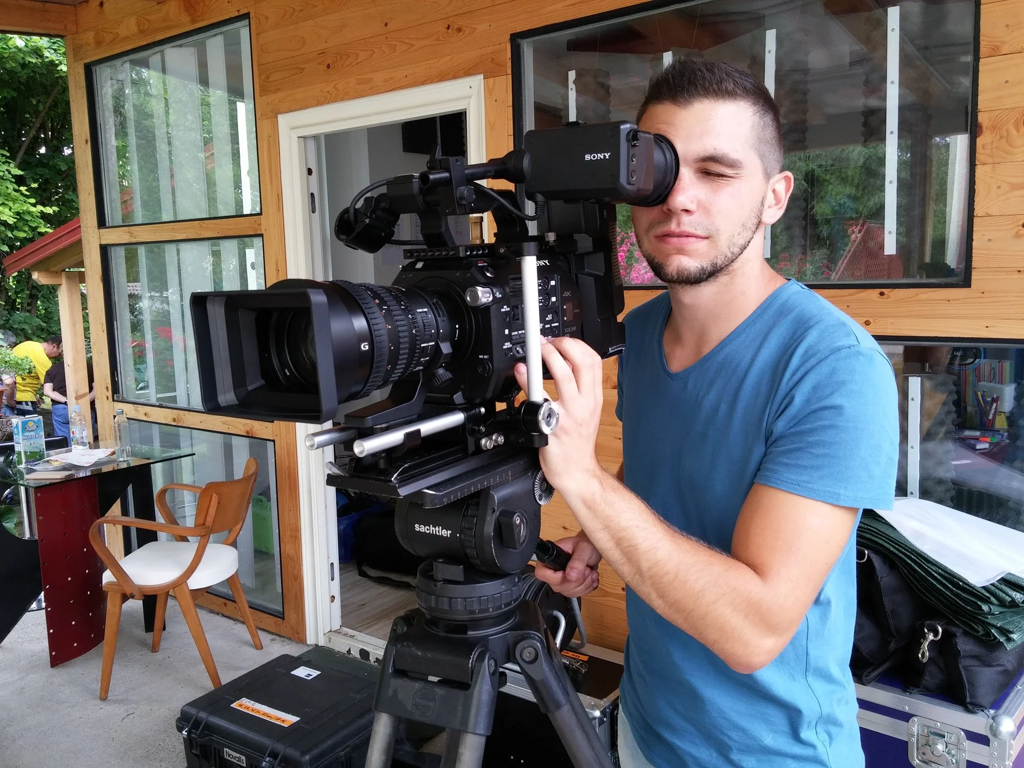 A man is operating a professional camera on a tripod indoors, with a wooden wall and large windows behind him.