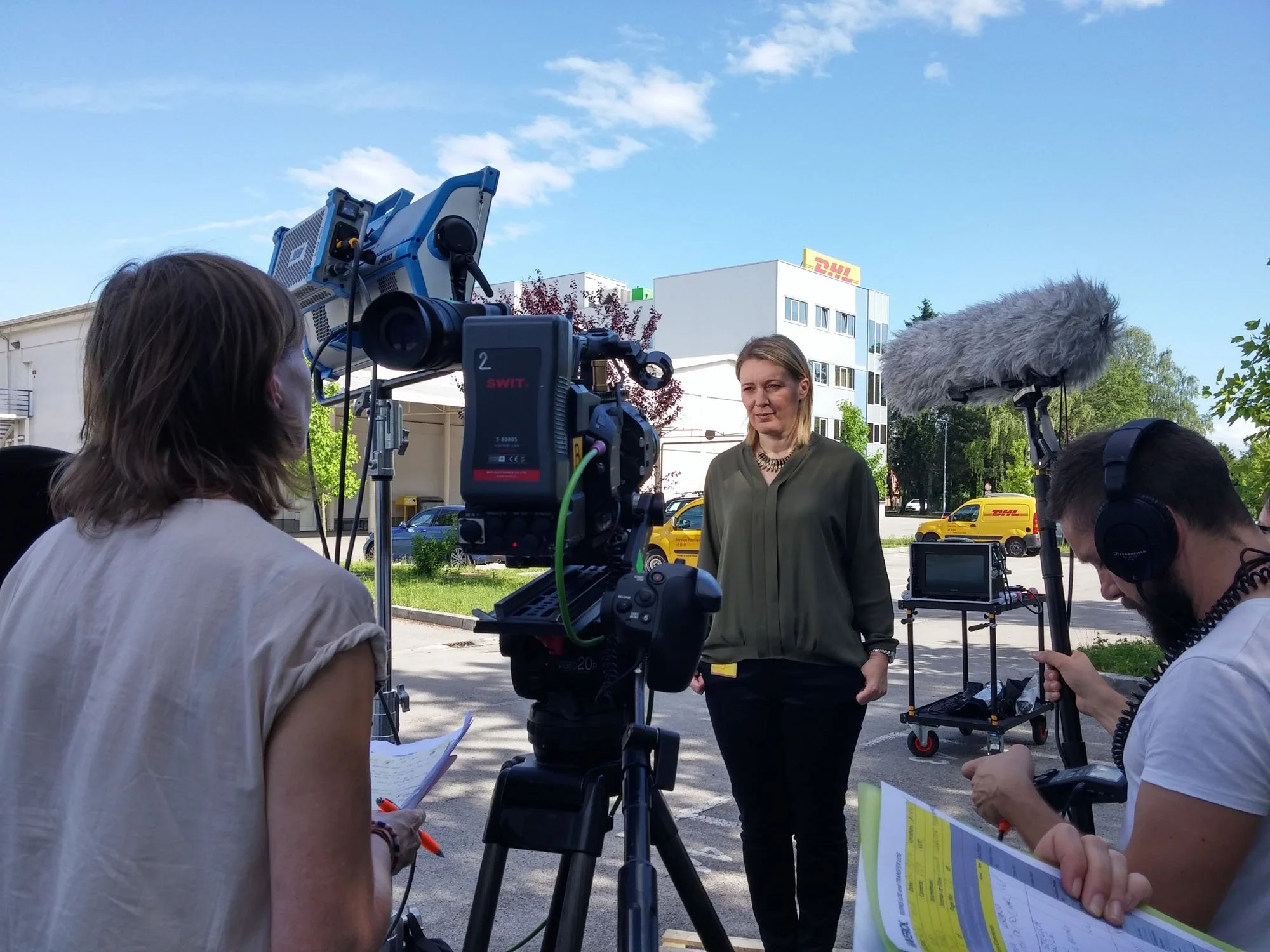 A woman is being interviewed outdoors by a small news crew with cameras, microphones, and notes, under a clear blue sky with some clouds, in front of white buildings and yellow DHL delivery vans.