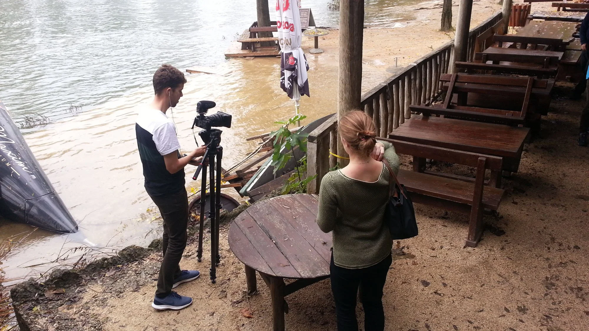 A young man filming with a camera on a tripod by a river, with a woman watching and a woman partially visible on the right, near a patio with empty wooden tables and chairs, during daytime.