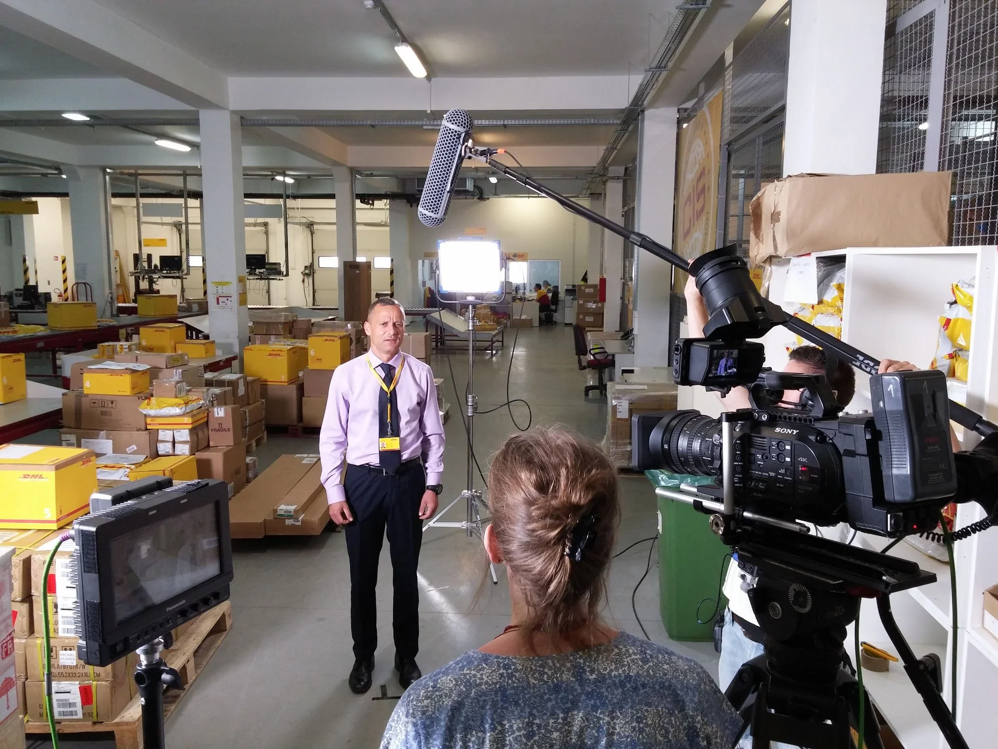 A man in a dress shirt and tie is being filmed in a warehouse with boxes and packages around. A camera crew is operating a large professional video camera and lighting equipment to record him.