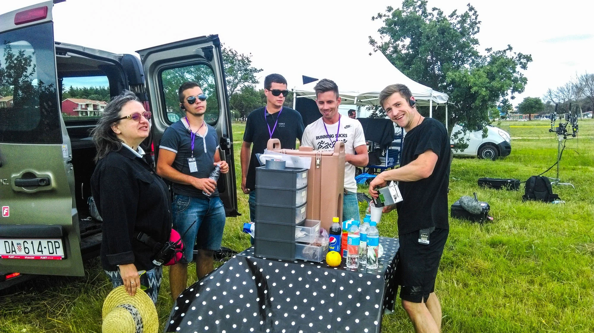 A group of five people standing around a table with snacks and drinks outdoors at a park, with equipment and a white tent in the background.