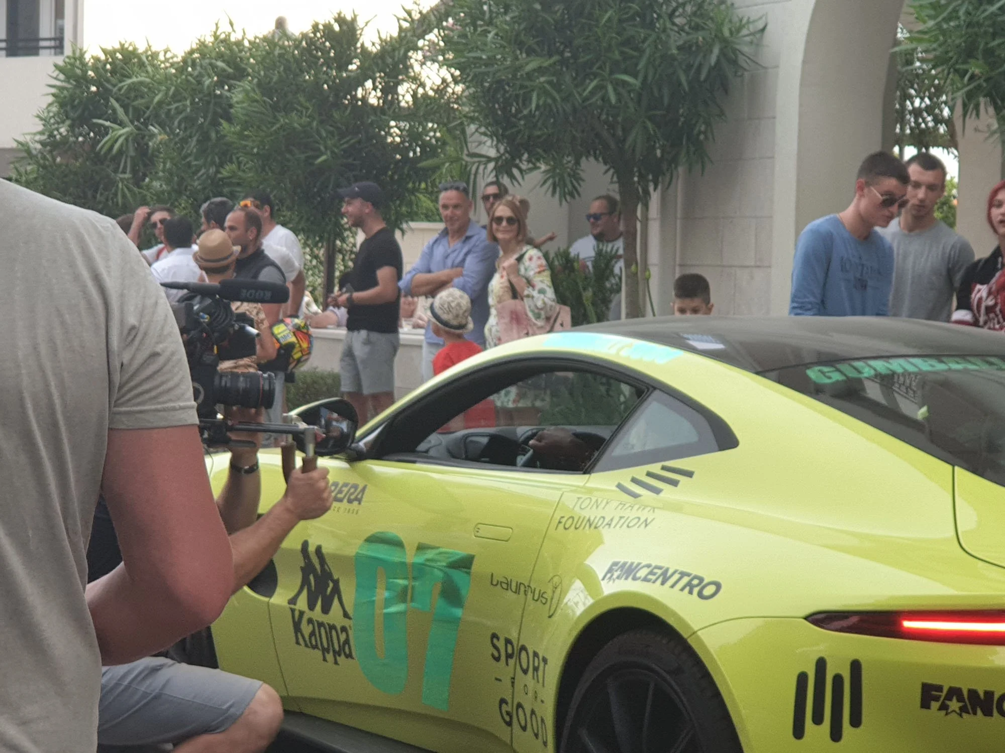 A group of people gather outdoors behind a yellow sports car with various logos and text on it, during what appears to be a public event or gathering.