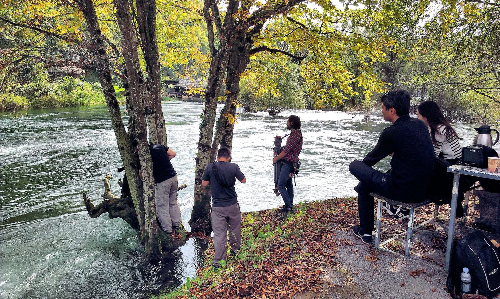A group of people by a river, surrounded by trees with green and yellow leaves. Some are standing near the water, while others are sitting on a bench. The scene appears to be in a park or natural setting with a small house or pavilion in the backgrou
