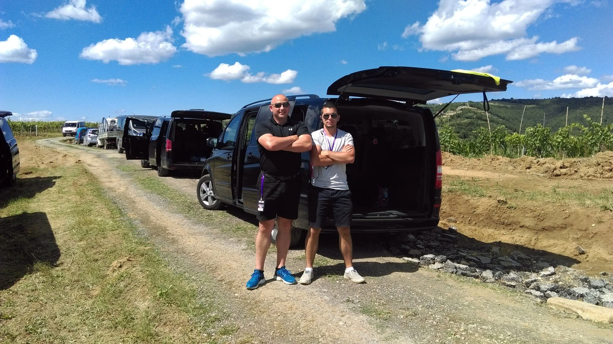 Two men standing with crossed arms behind a black SUV in a rural area with a line of parked cars and a vineyard in the background under a partly cloudy sky.