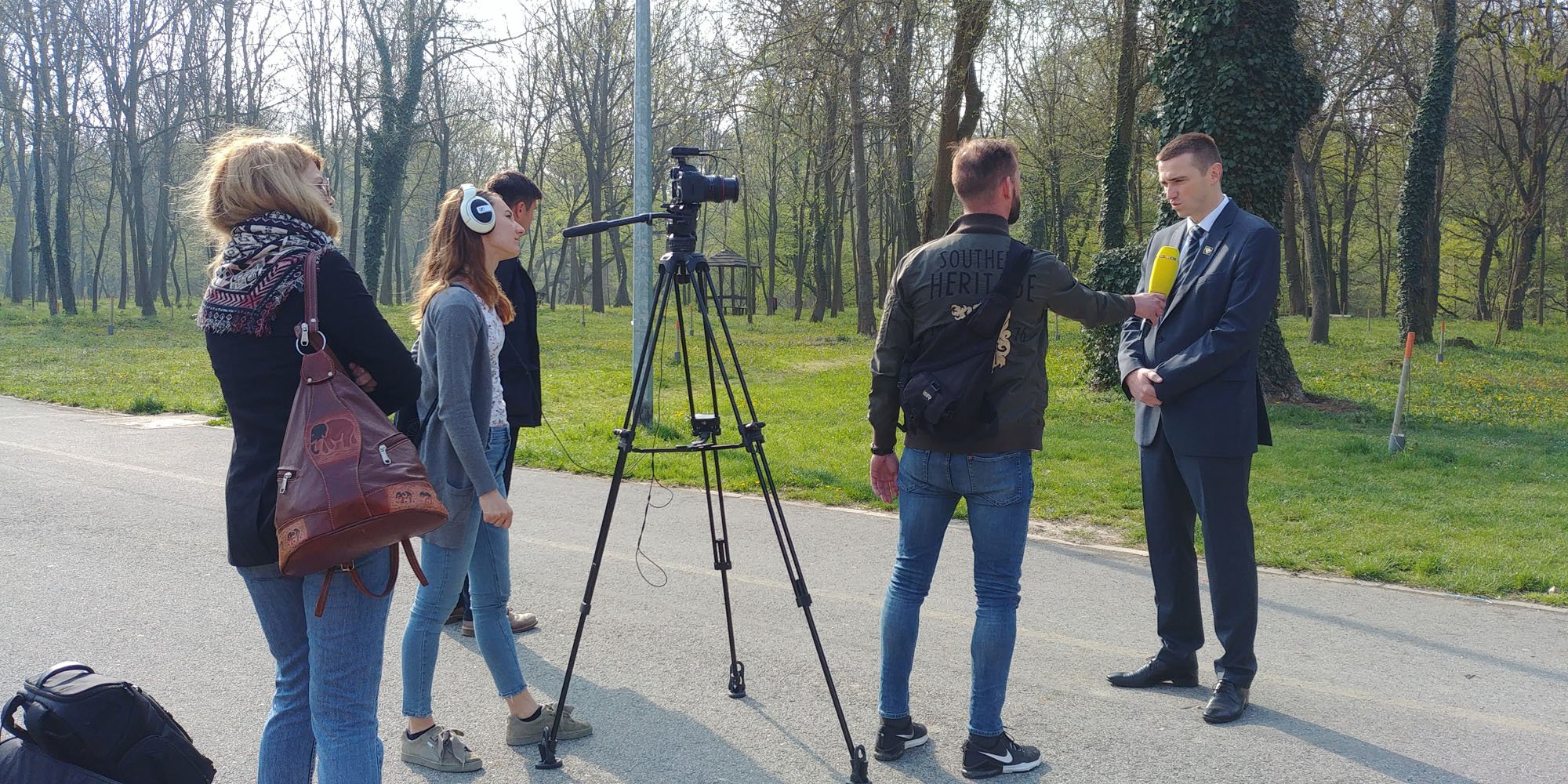 A group of people filming an interview outdoors on a paved path in a park with trees and grass in the background. One person is being interviewed with a yellow microphone, while others observe. A camera on a tripod is pointed at the interviewee.