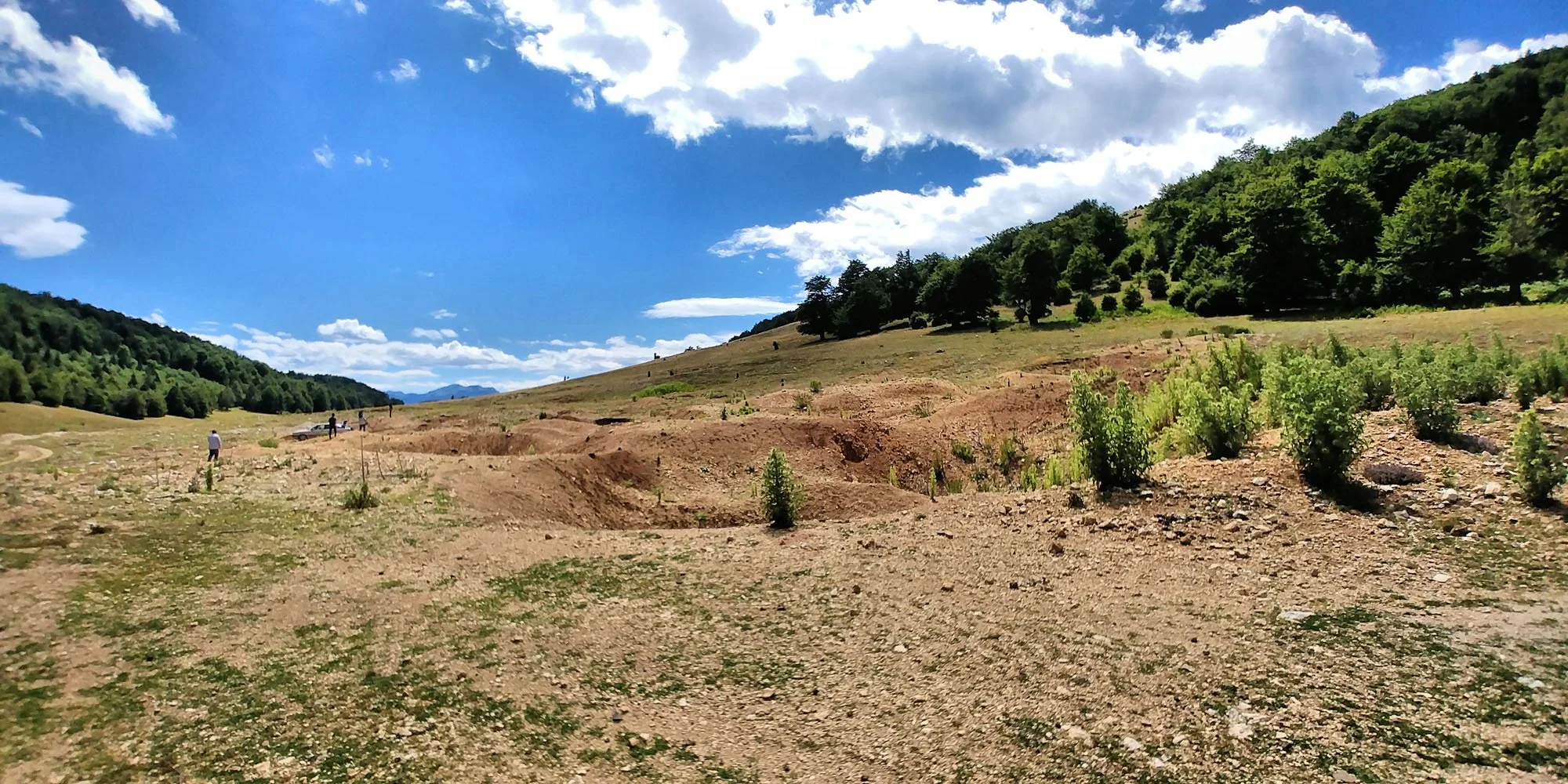 A hilly landscape with sparse vegetation, small trees, and people walking near parked cars under a partly cloudy sky.