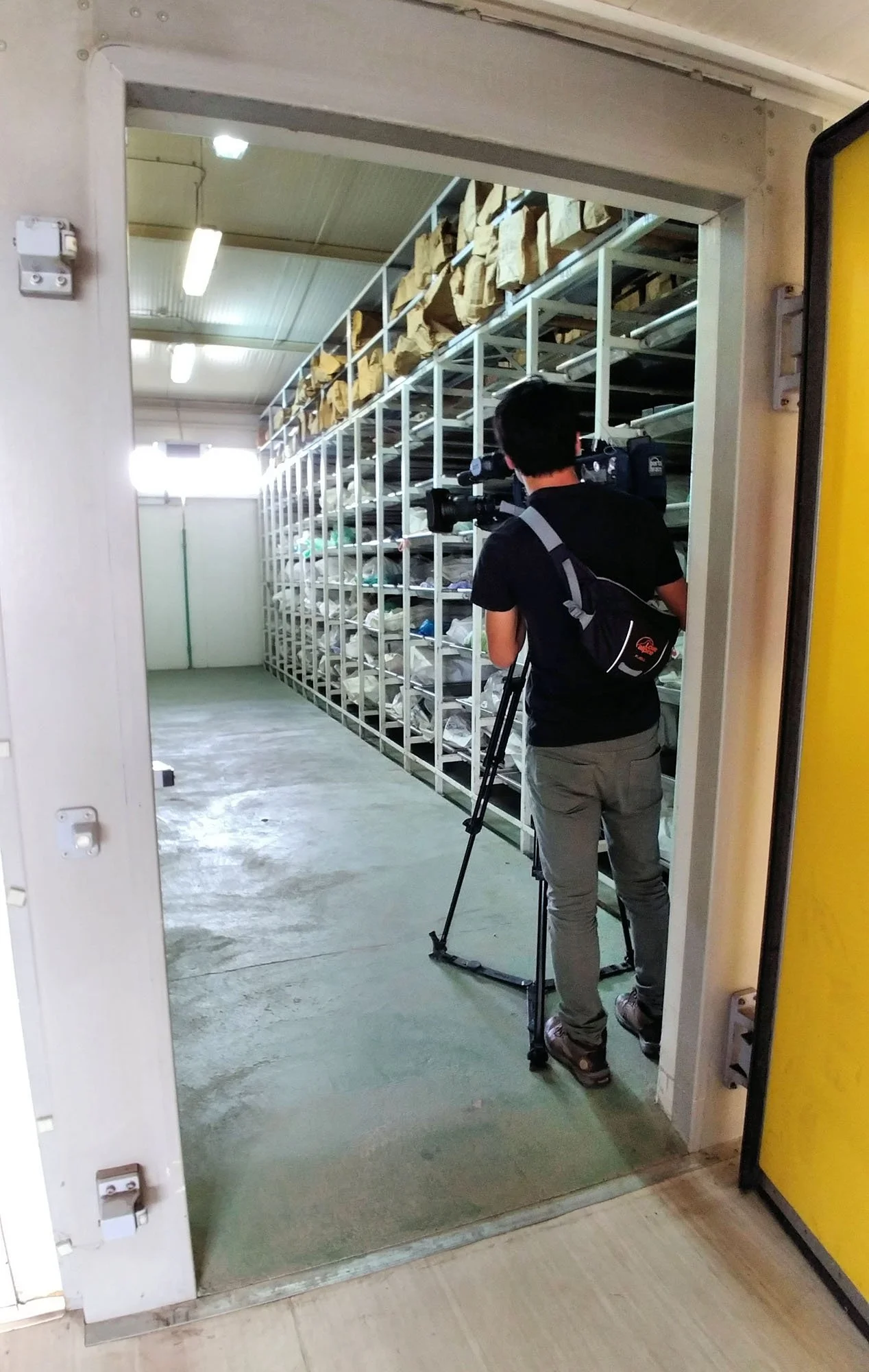 A person operating a camera on a tripod capturing footage of shelves filled with brown paper packages in a storage room or warehouse.