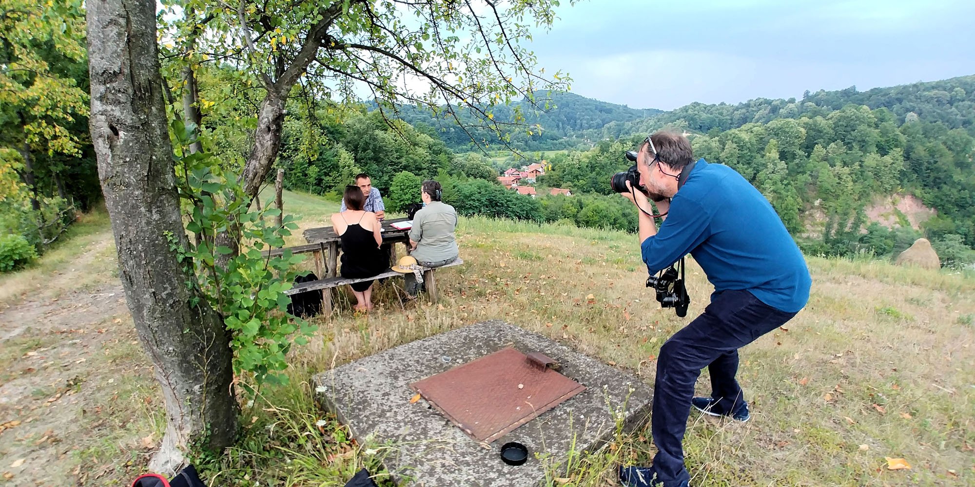 Photographer taking a picture of three people sitting at a picnic table outdoors in a rural, hilly landscape with trees and houses in the background.