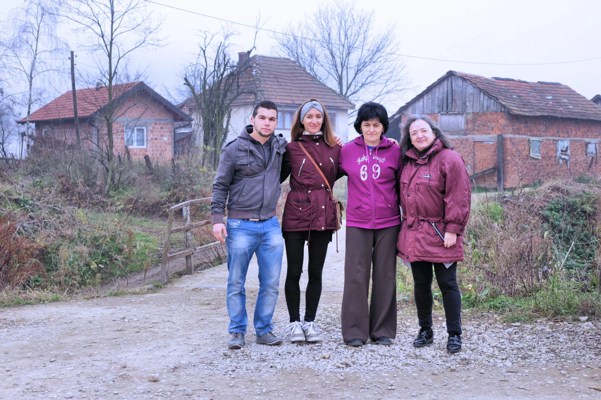 Four women and one man standing on a dirt path outdoors, with houses and leafless trees in the background.