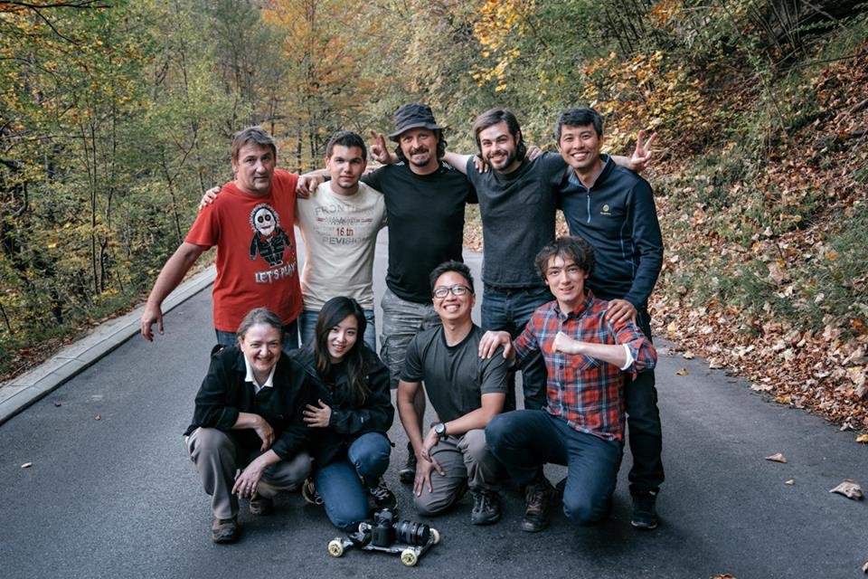 A group of nine friends posing on a paved road in a wooded area during fall, some kneeling and others standing, smiling and making peace signs, with a camera on the ground in front.