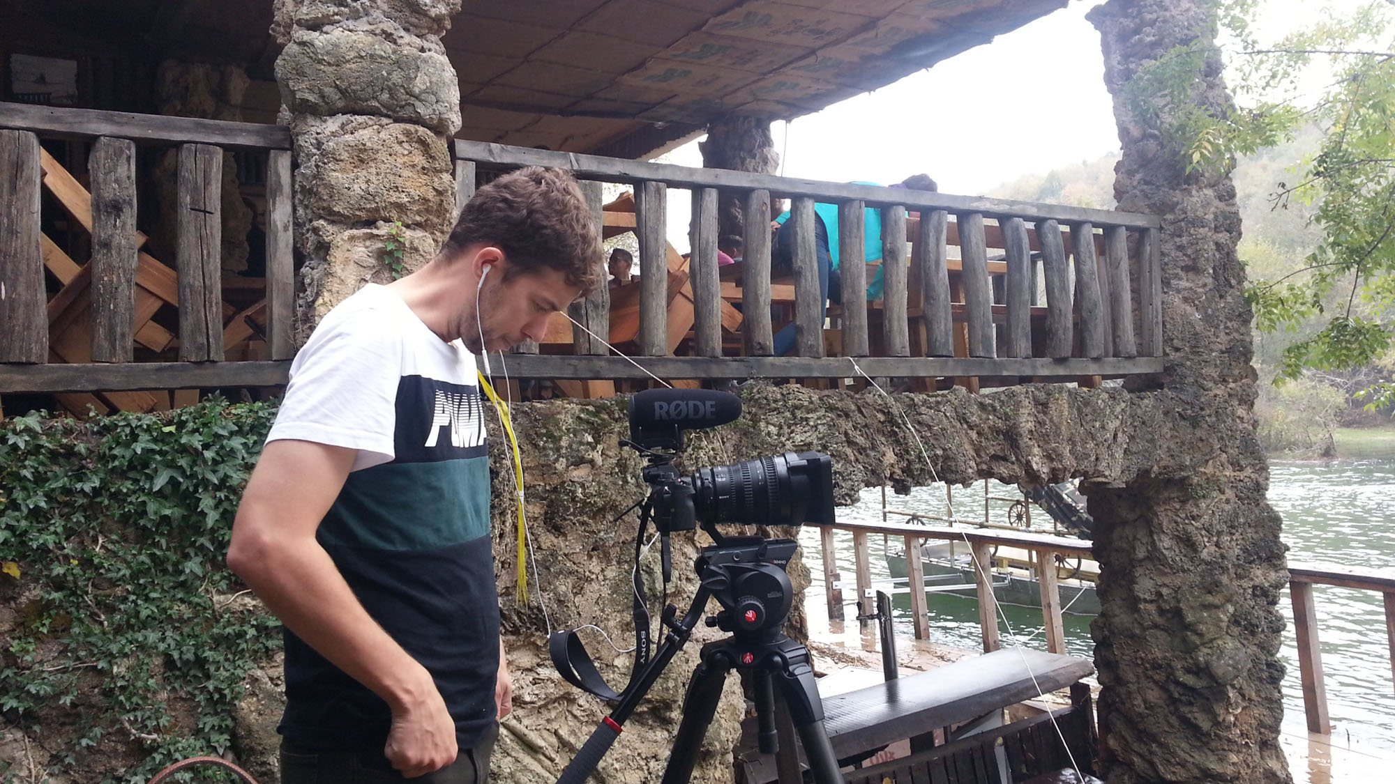 A man with a camera recording setup, wearing earphones, standing near a rocky riverbank with a wooden bridge and several people on the bridge in the background.